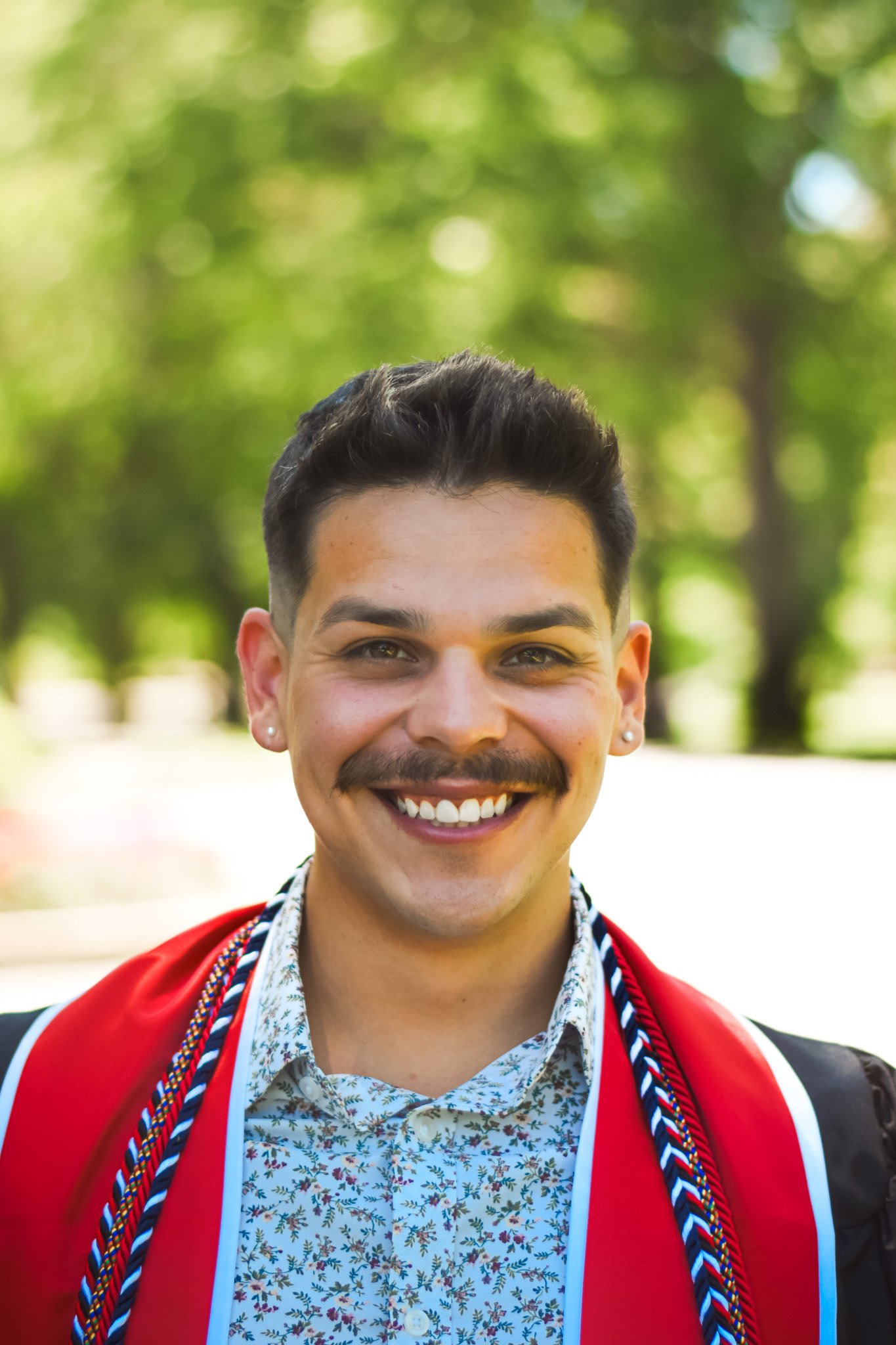 Close-up of a smiling person with short dark hair, mustache, and earrings, wearing a floral shirt and a red graduation gown with cords, outdoors with blurry green trees in the background.