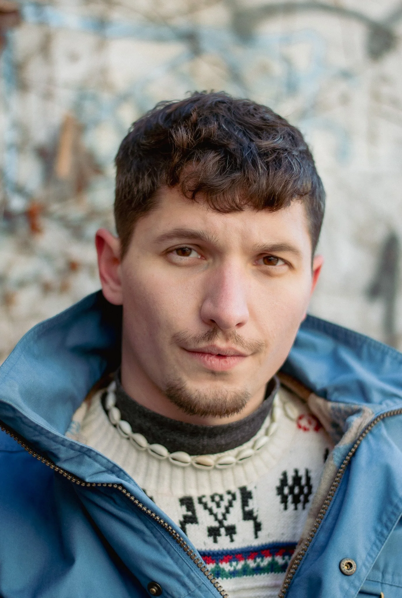 Close-up of a young man with brown hair, brown eyes, wearing a blue jacket and a sweater with a Christmas pattern, outdoors in a natural setting.