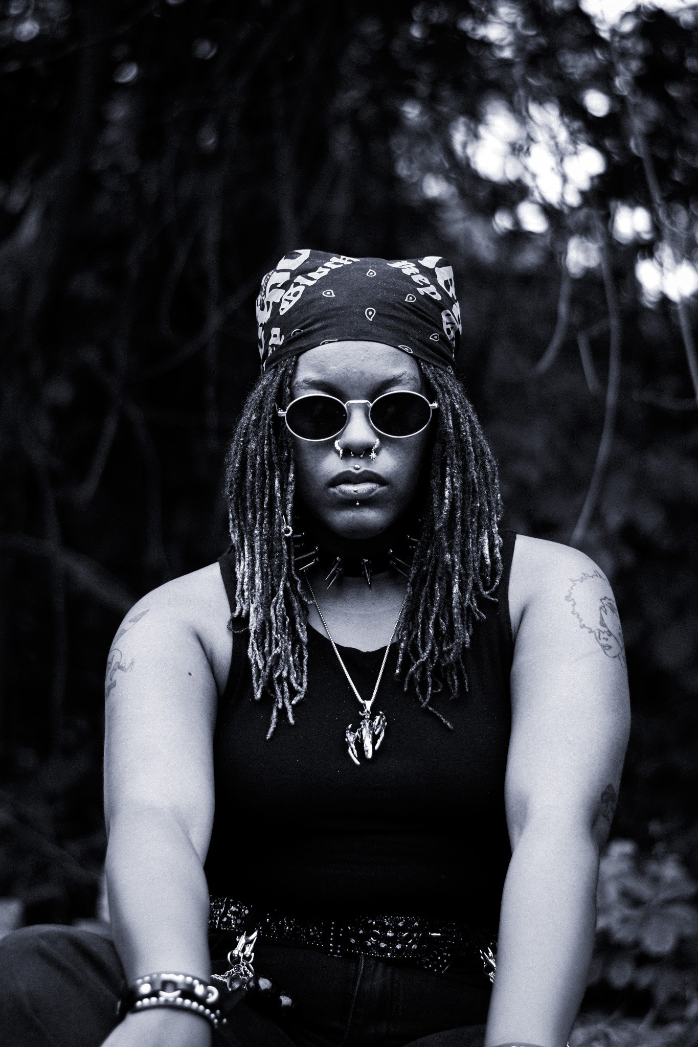 Black and white portrait of a young woman with dreadlocks wearing round sunglasses, a bandana, and multiple necklaces, sitting outdoors in a natural setting.