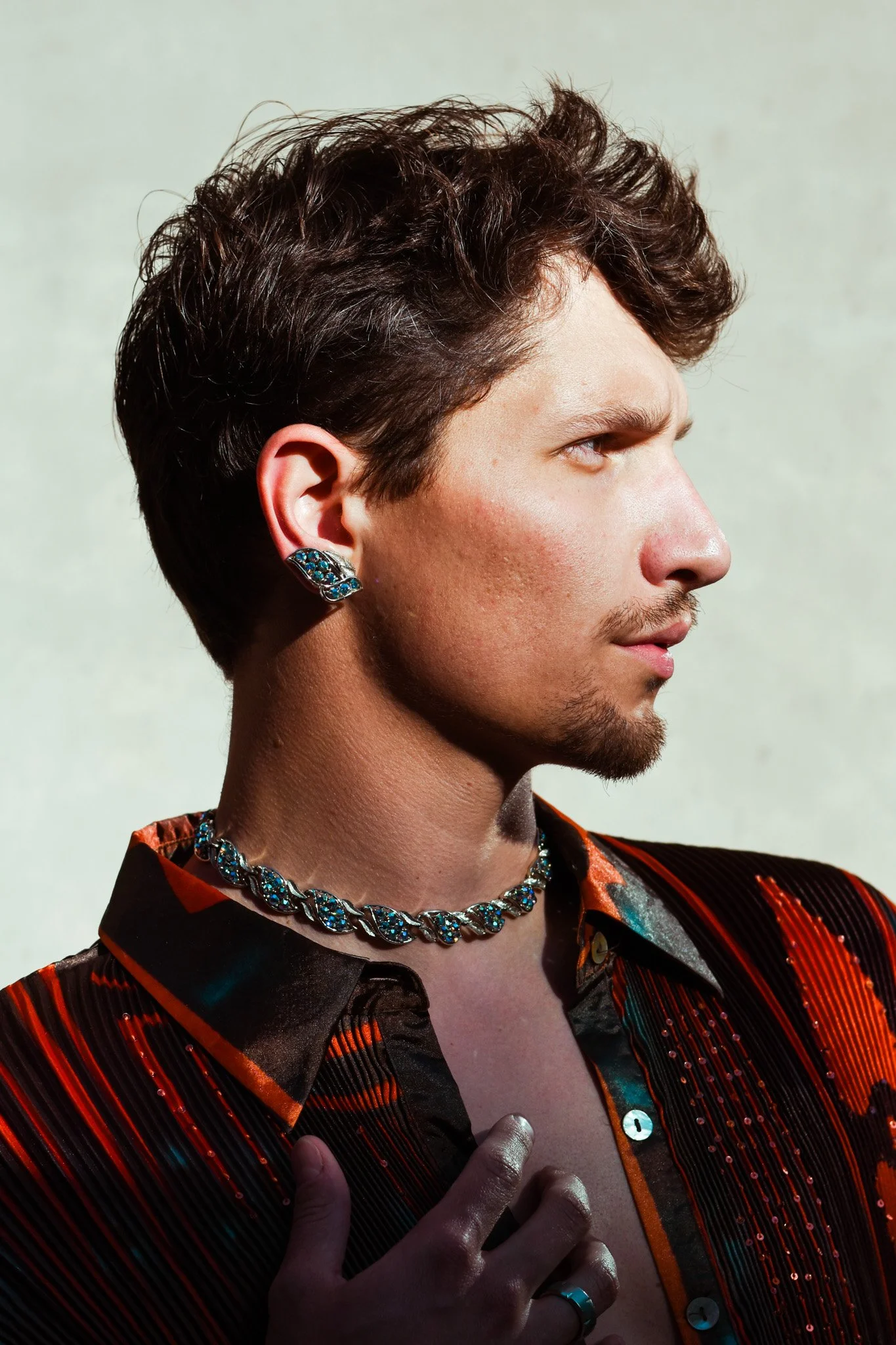 A young man with brown curly hair and facial hair wearing a striped, colorful shirt, a ring on his finger, a turquoise ring on his thumb, earrings, and a necklace, with his hand on his chest and his face turned to the right.
