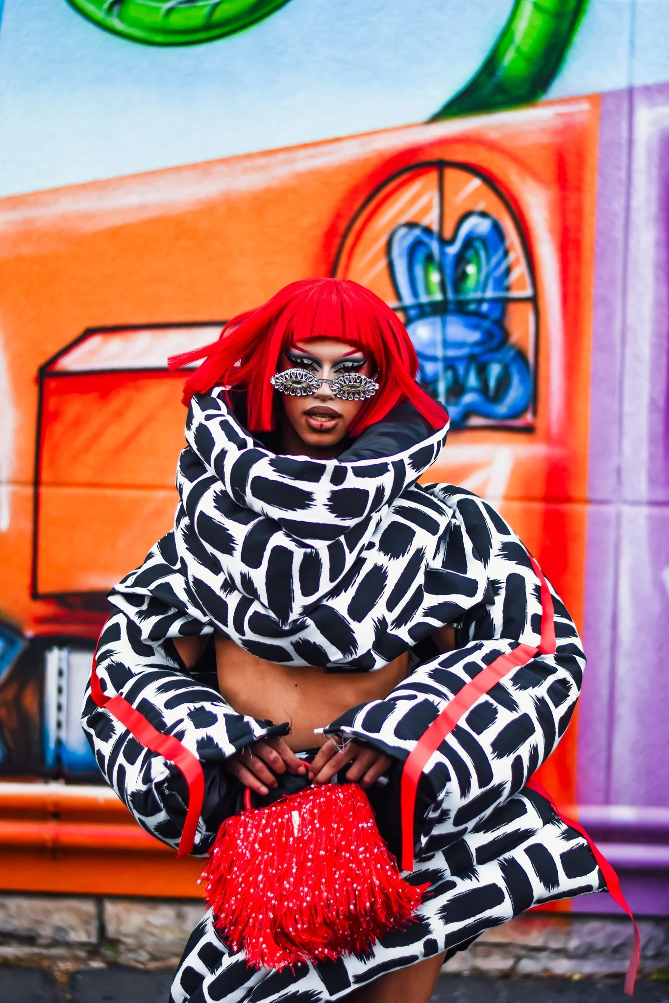 A person with bright red hair and patterned glasses poses in front of colorful graffiti art, wearing a matching black and white patterned outfit with red accents, holding a red fringed purse.
