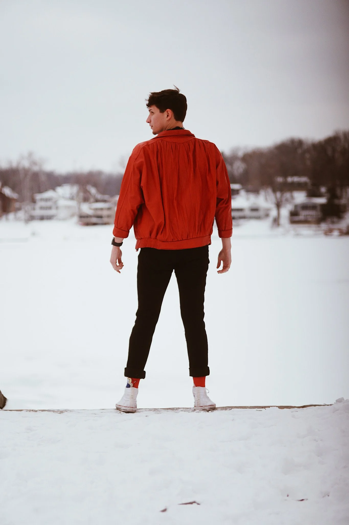 A young man in a red jacket and white sneakers standing on snow-covered ground, facing sideways with a winter landscape and trees in the background.