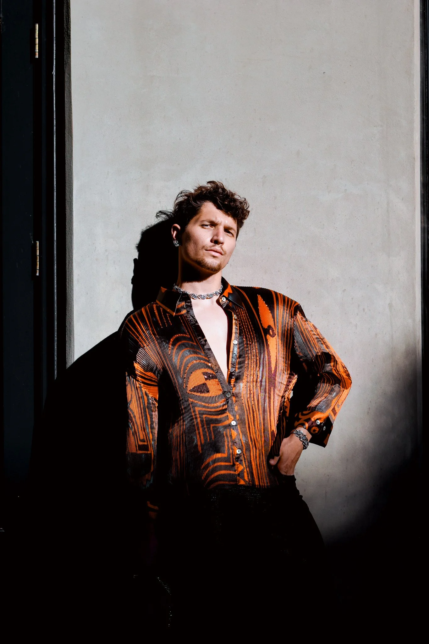 A man with curly hair and a goatee posing with one hand on his hip against a plain wall, wearing a vibrant patterned orange and black shirt, necklace, earrings, and bracelets.