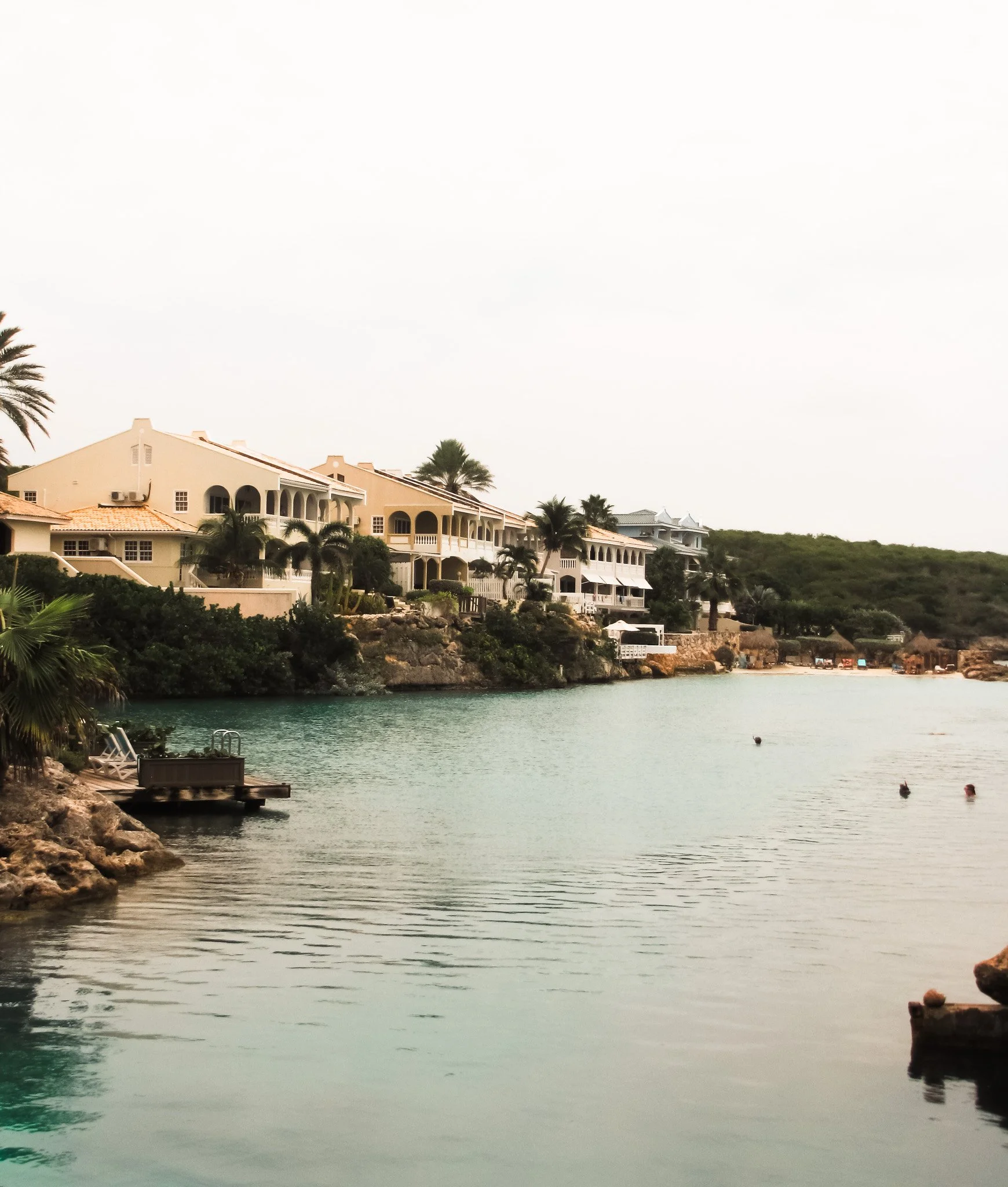 Waterside scene with calm water, large white buildings with arches and balconies, palm trees, and a rocky shoreline, possibly a resort or hotel near a beach.
