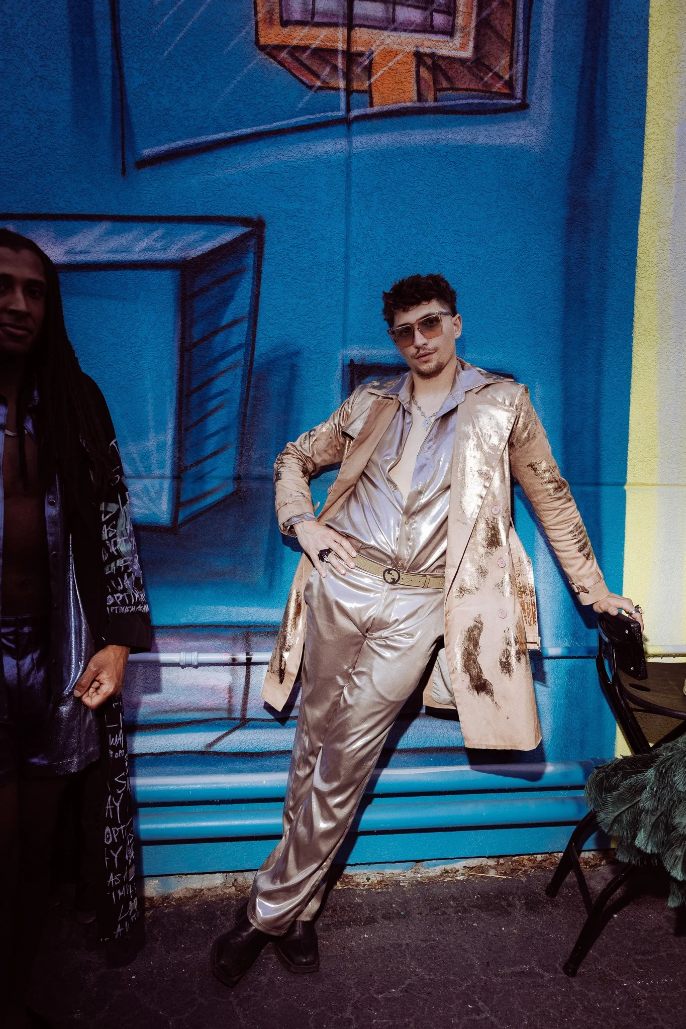 A man wearing a shiny gold suit with a matching trench coat poses in front of a colorful wall with abstract geometric artwork. He is wearing sunglasses, jewelry, and holding a black stroller. A part of another person is visible on the left side of th