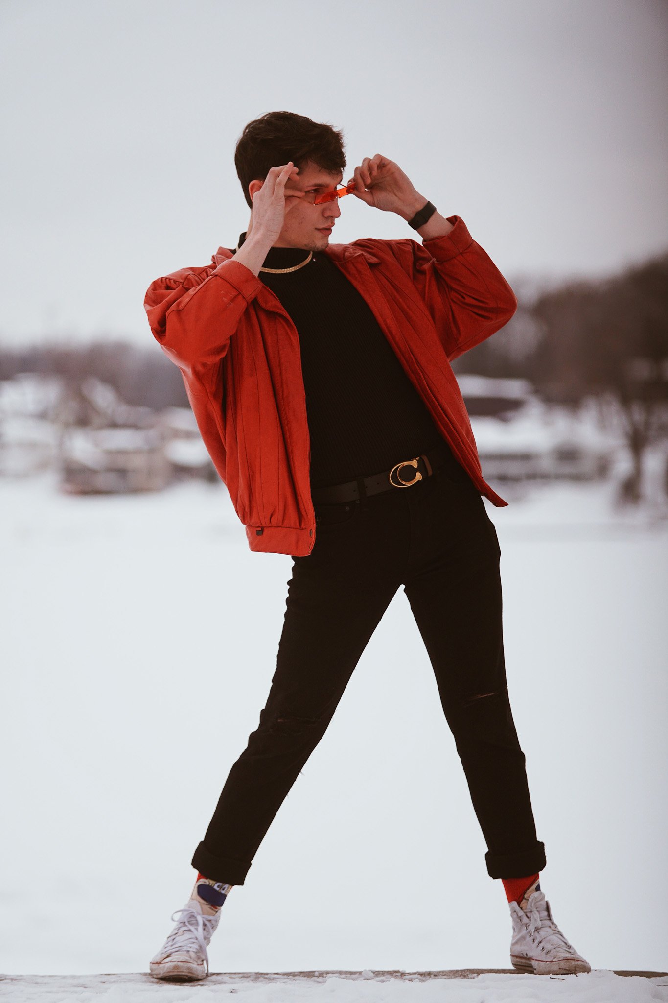 A young man in a red jacket, black shirt, and black pants standing in a snowy outdoor setting, adjusting orange sunglasses, with snow and trees in the background.