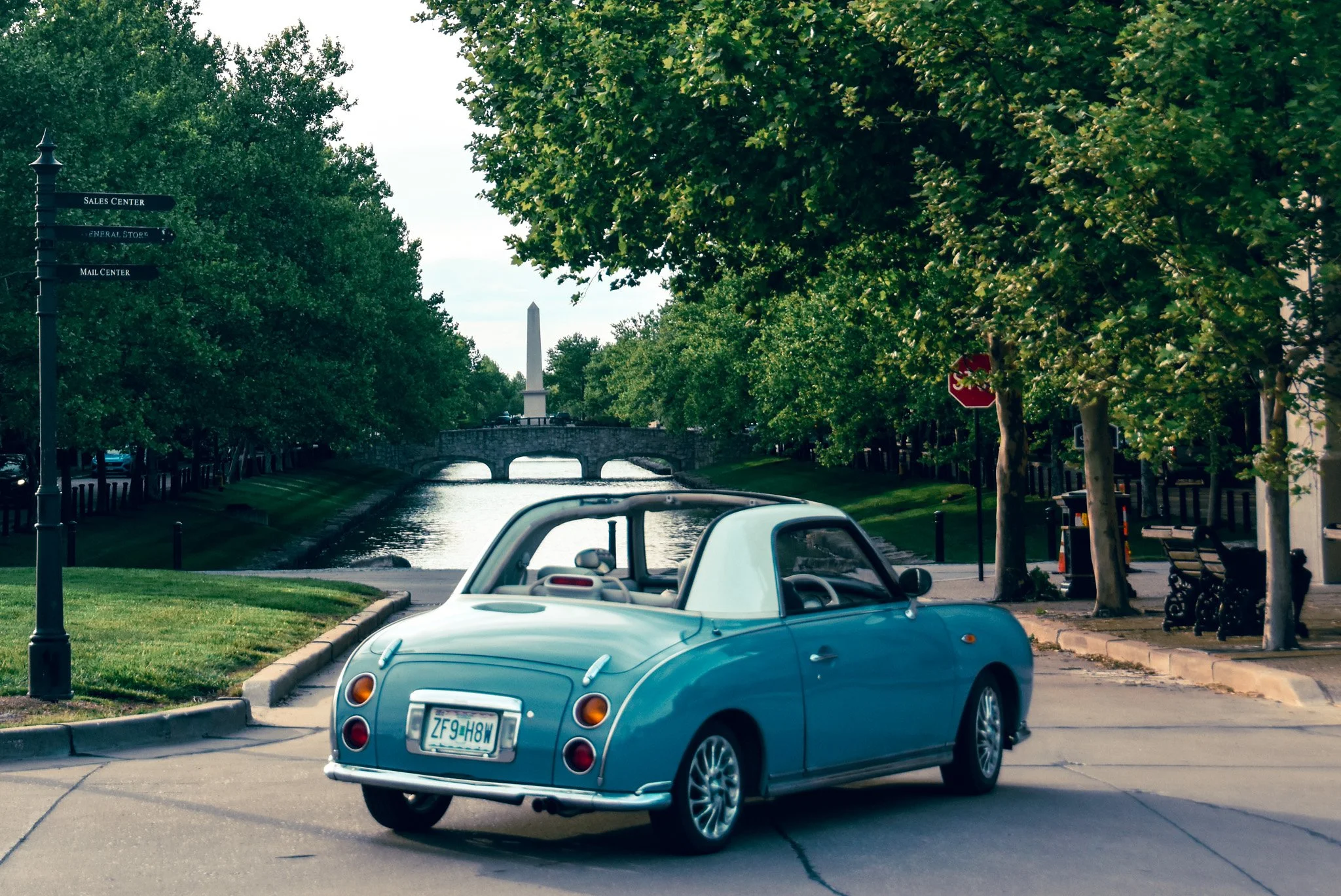 A light blue vintage-style convertible car with a white interior parked on a park sidewalk near a canal. The canal has a stone bridge and an obelisk monument in the background. The surroundings are green with trees lining the street, and there are be