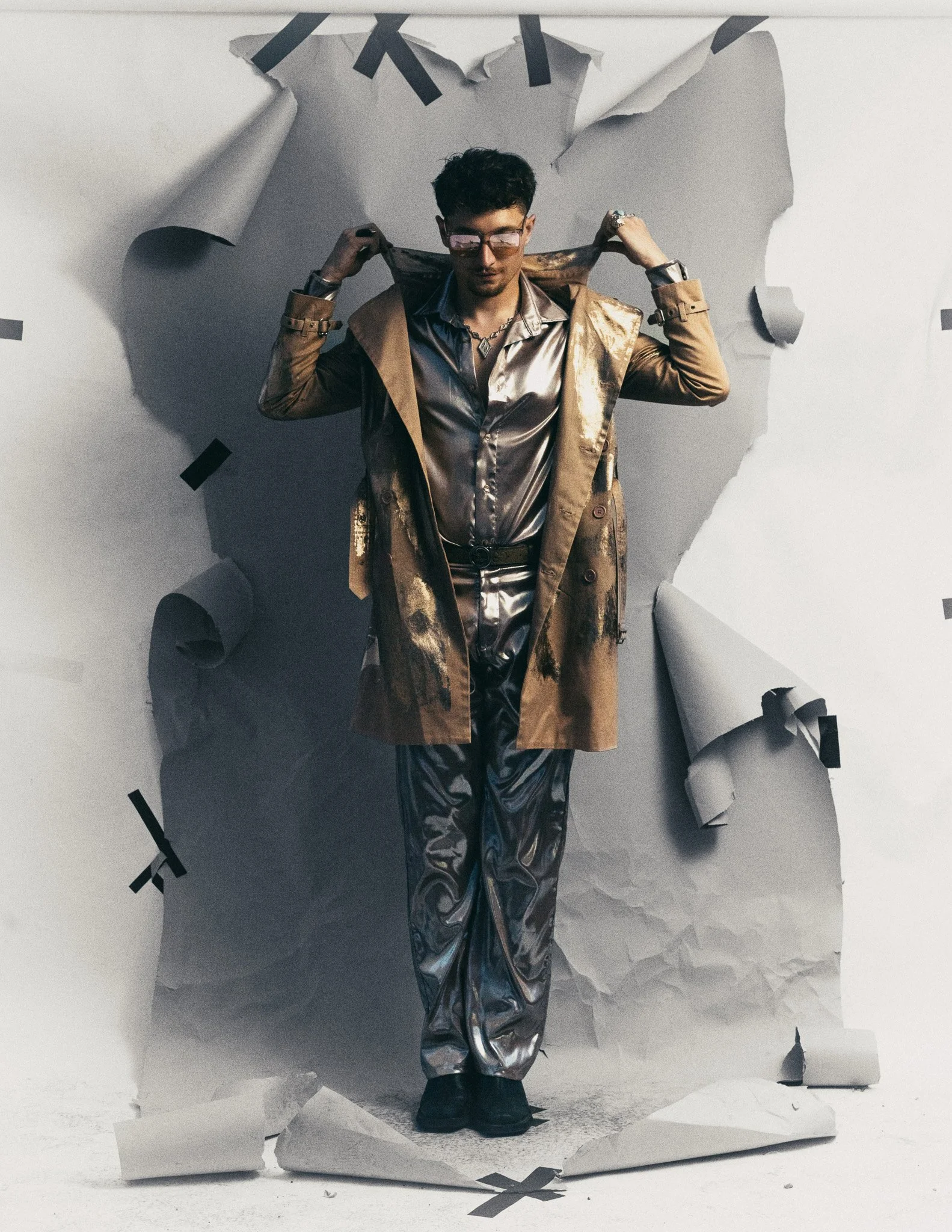 A young man in shiny metallic clothing and sunglasses standing against a torn paper backdrop, holding a jacket over his shoulders and striking a confident pose.