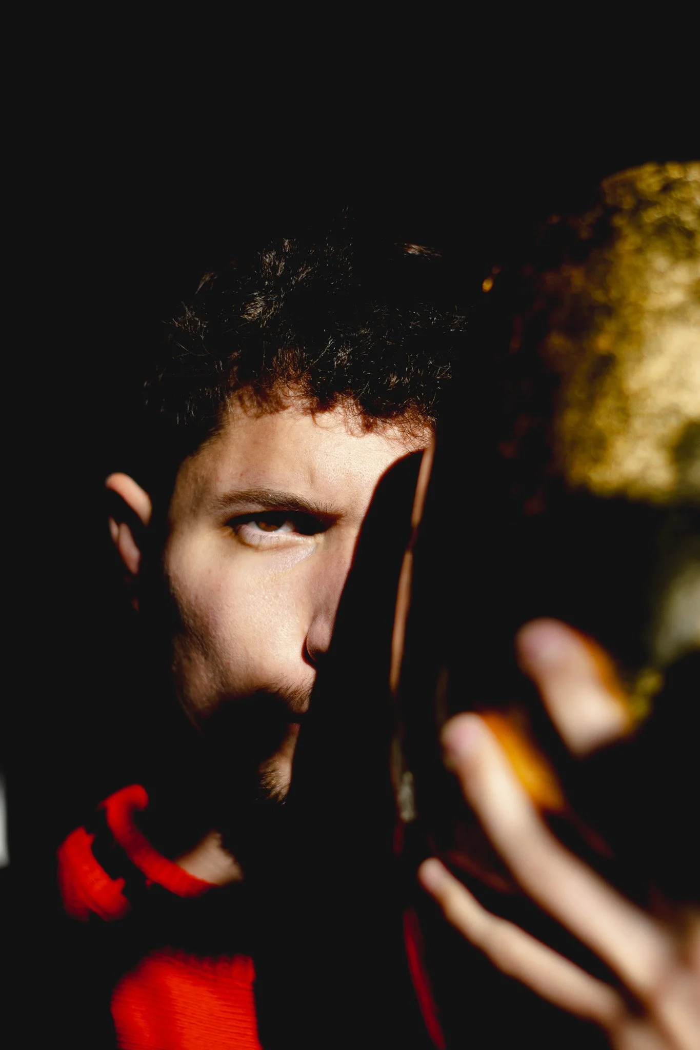 A close-up of a young man with dark, curly hair, wearing a black and red hoodie, looking at the camera with a serious expression and his hand partially covering his face, against a black background with some gold and red highlights.