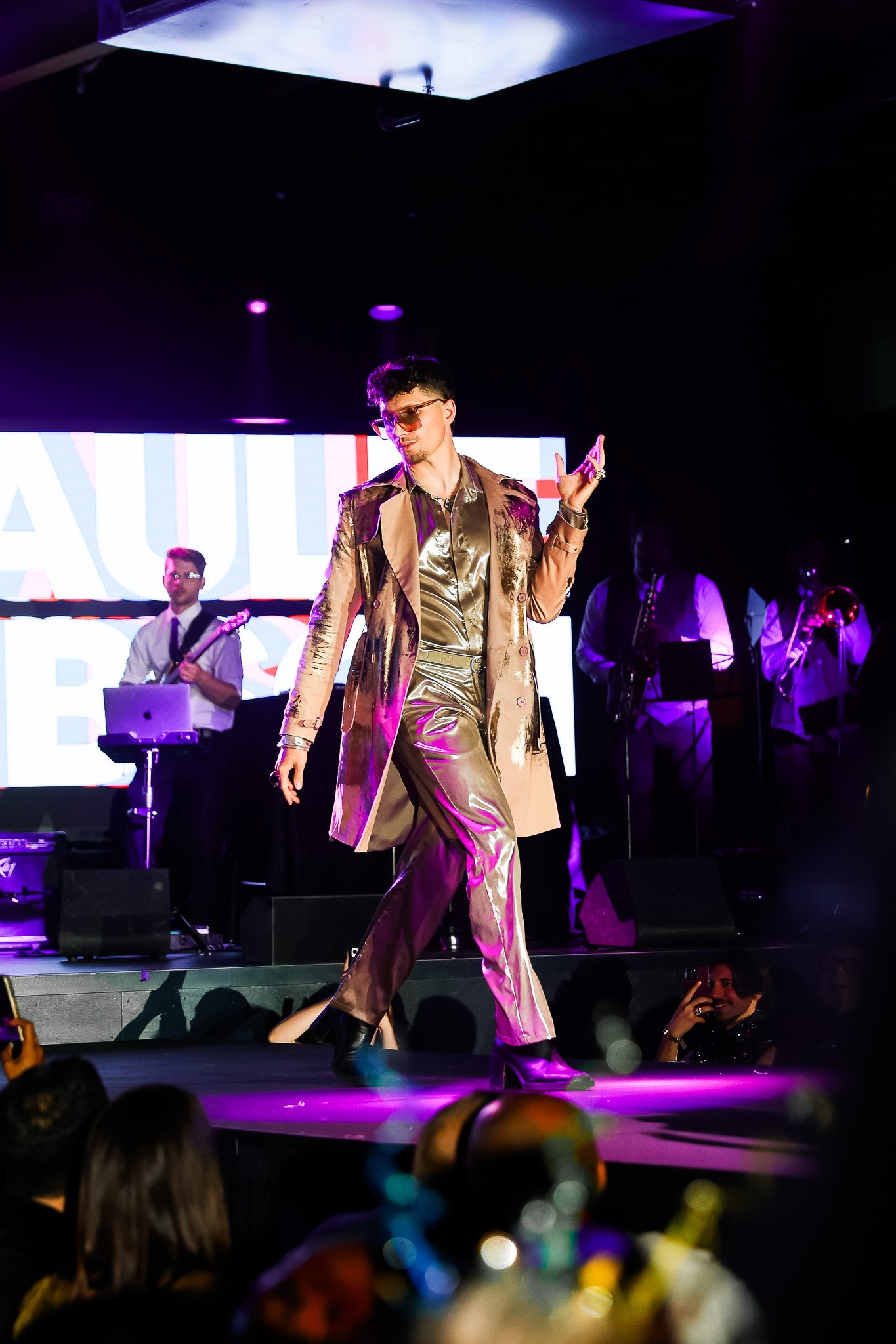 A man dressed in shiny gold clothes and sunglasses performing on stage with a band in the background. The stage has purple lighting and a large screen behind him displays text.