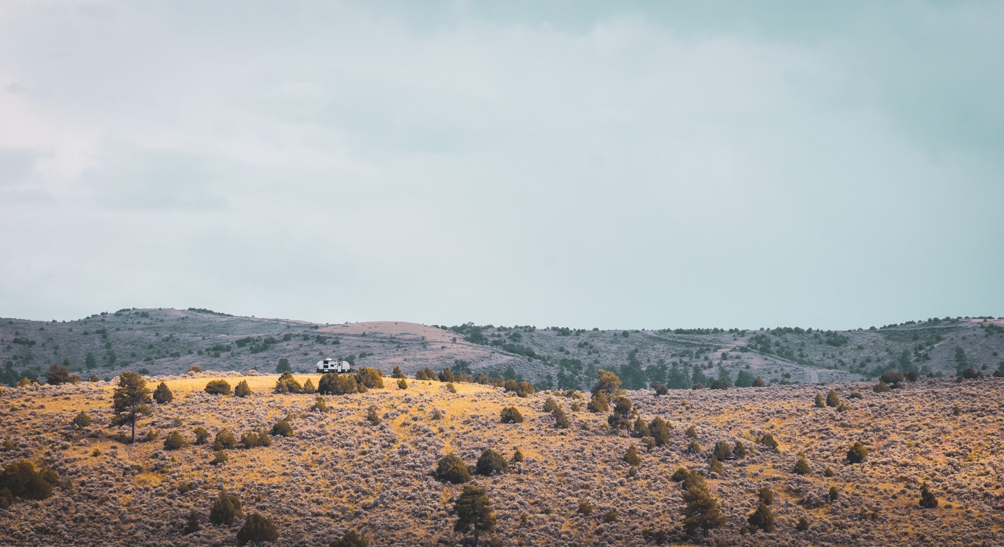 A landscape with a rolling hill covered in dry grass and bushes, a white camper van on the hill, and a cloudy sky overhead.