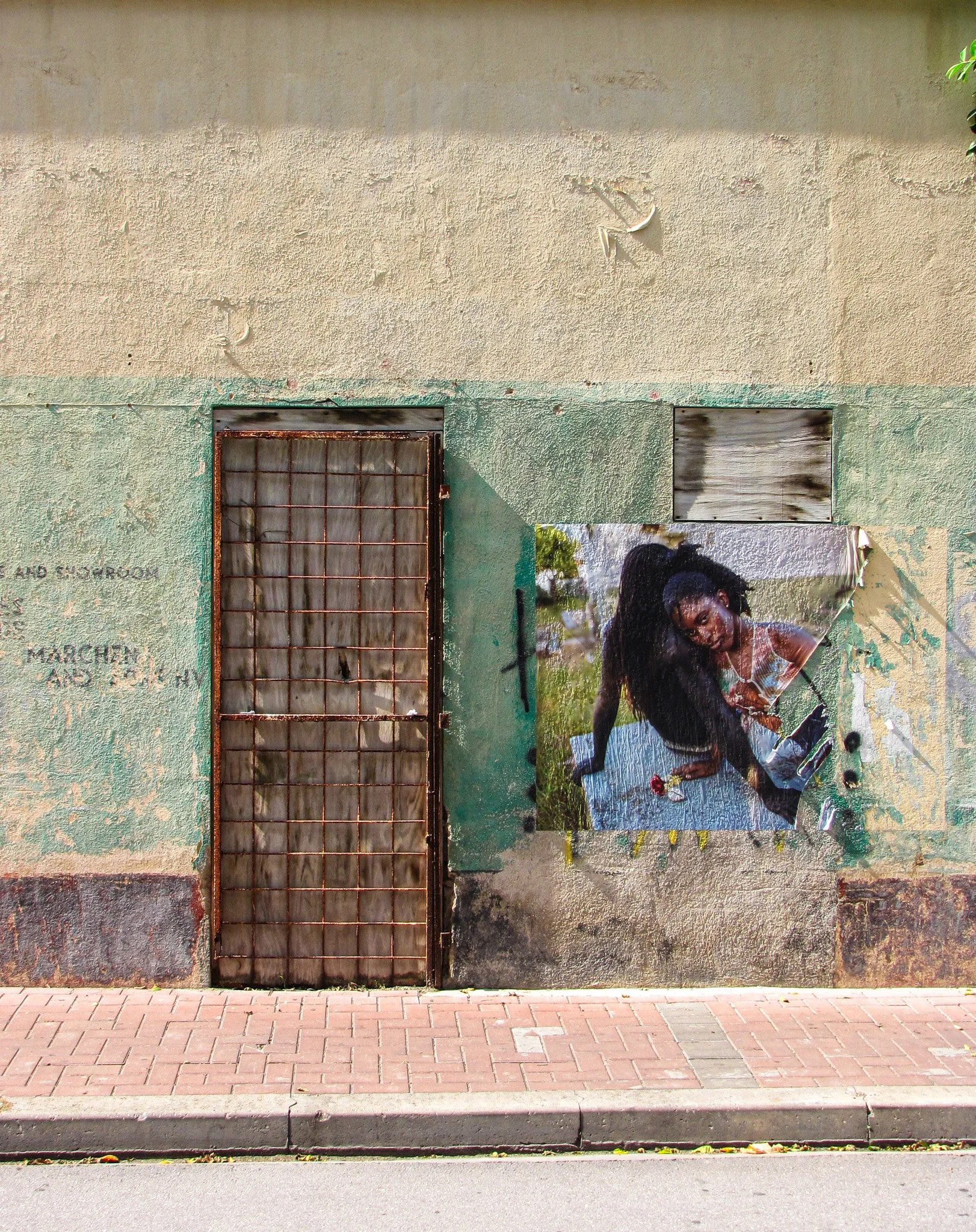 A beige and green textured exterior wall with a rusty metal grid door, a wooden boarded-up window, and a large mural of a woman with long dark hair and a light-colored top, holding a flower, with green foliage in the background.