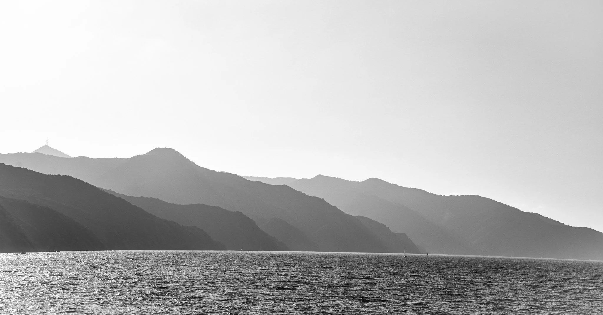 Black and white photo of a body of water with mountain ranges in the background and a sailboat on the water.