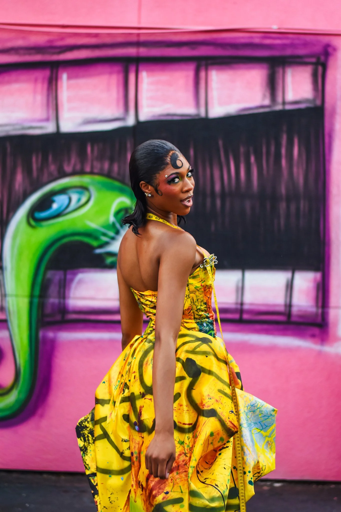 A woman with colorful makeup and a yellow tie-dye dress sitting in front of a vibrant graffiti wall.