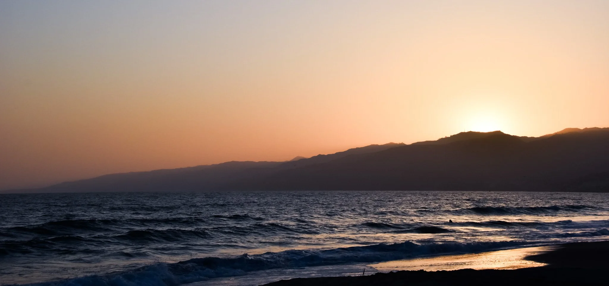 Sun setting behind distant mountains over ocean, with waves crashing on the shore in the foreground.