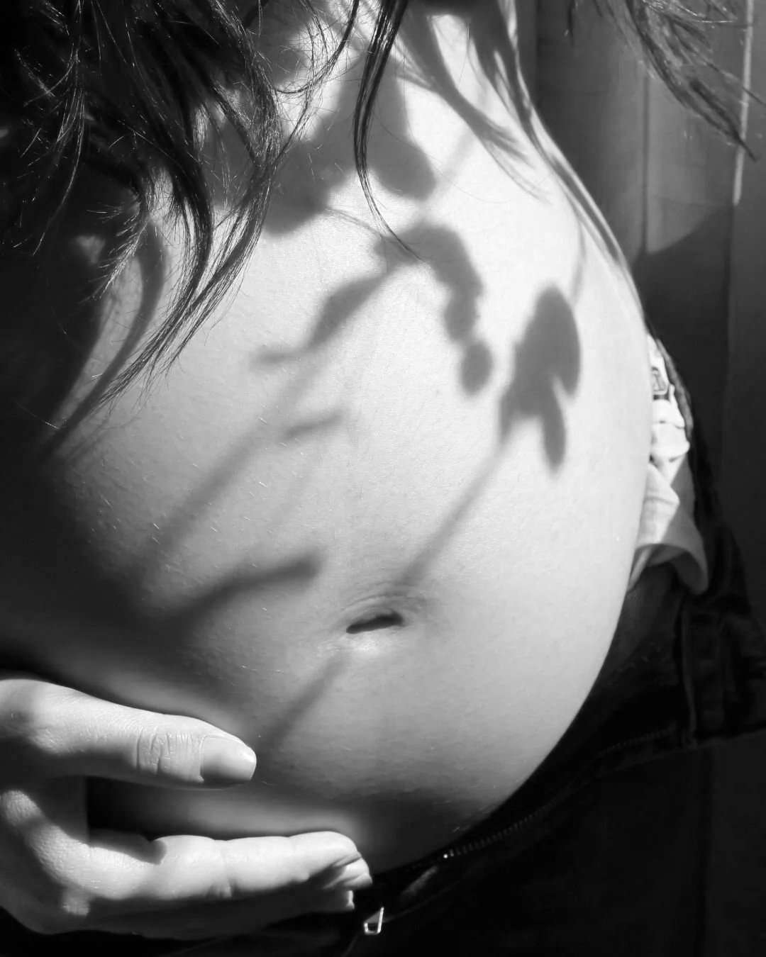 Black-and-white portrait of a pregnant woman's belly with natural leaf shadows, representing pregnancy, maternal care, and personalized women's health at every stage of life.