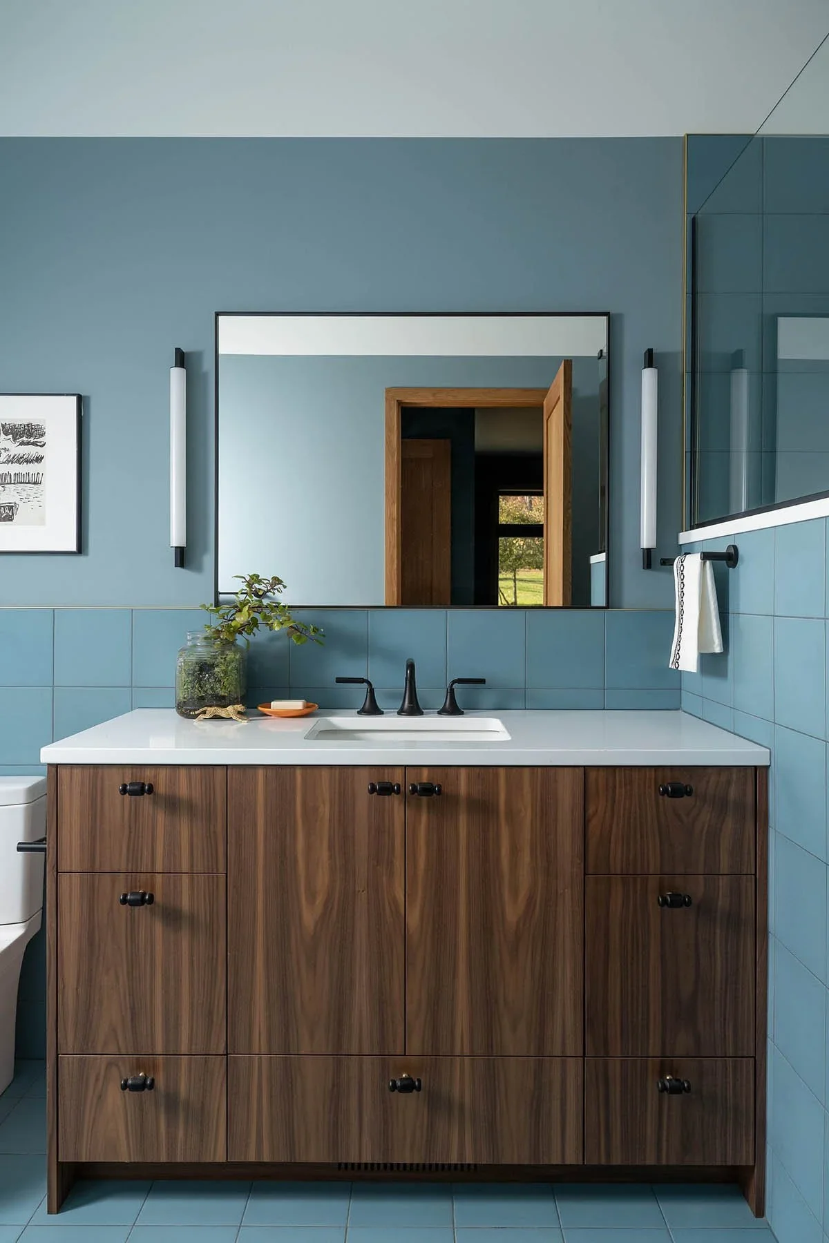 Modern bathroom with blue tiled walls, a large mirror above a white countertop with a black faucet, a wooden vanity with black knobs, a plant in a glass jar, and decor. Two elongated wall lights flank the mirror, and a towel hangs on a black rod.