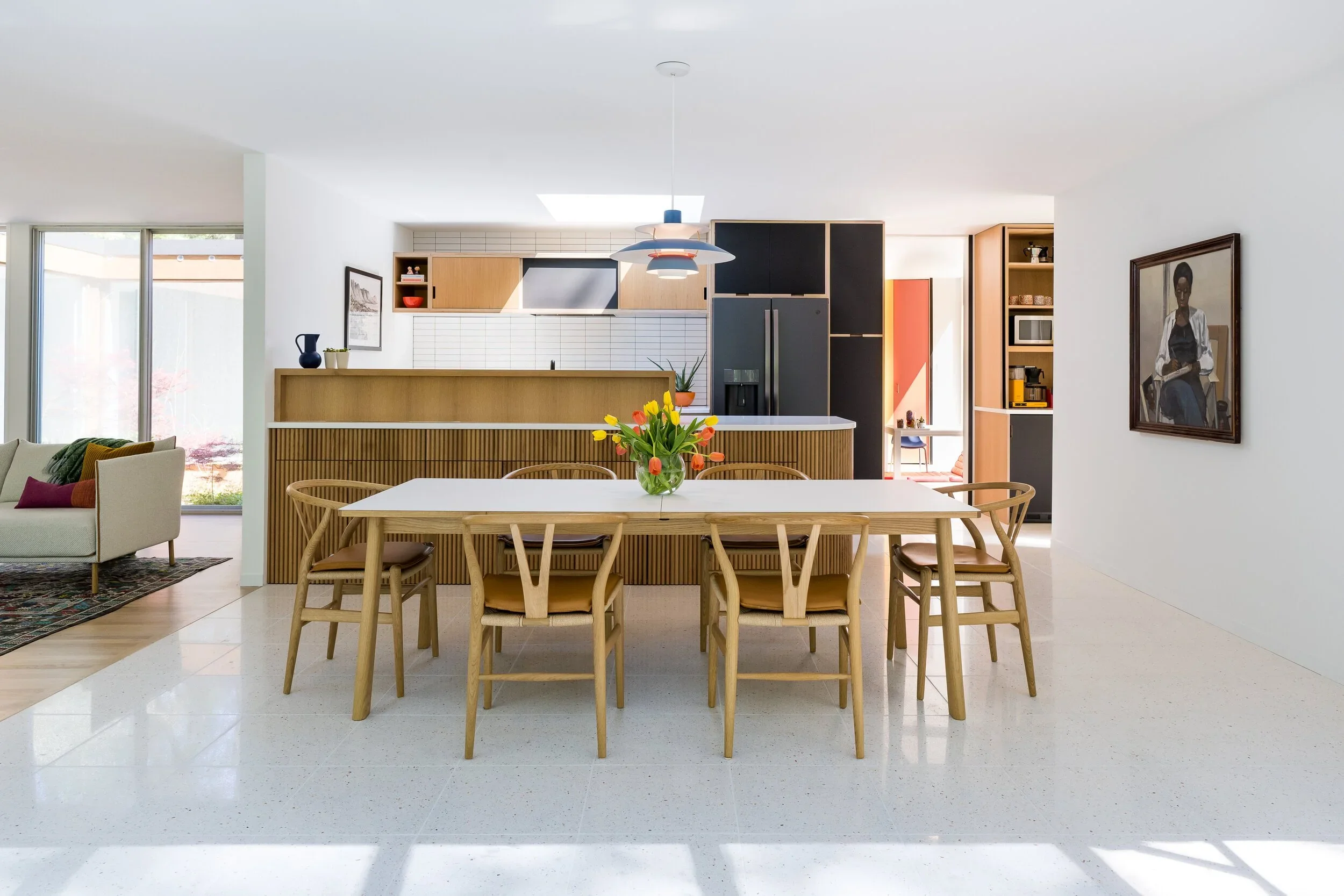 Open-concept kitchen and dining area with a wooden table and chairs, a vase of tulips, built-in shelving with kitchen appliances, and a portrait on the wall.