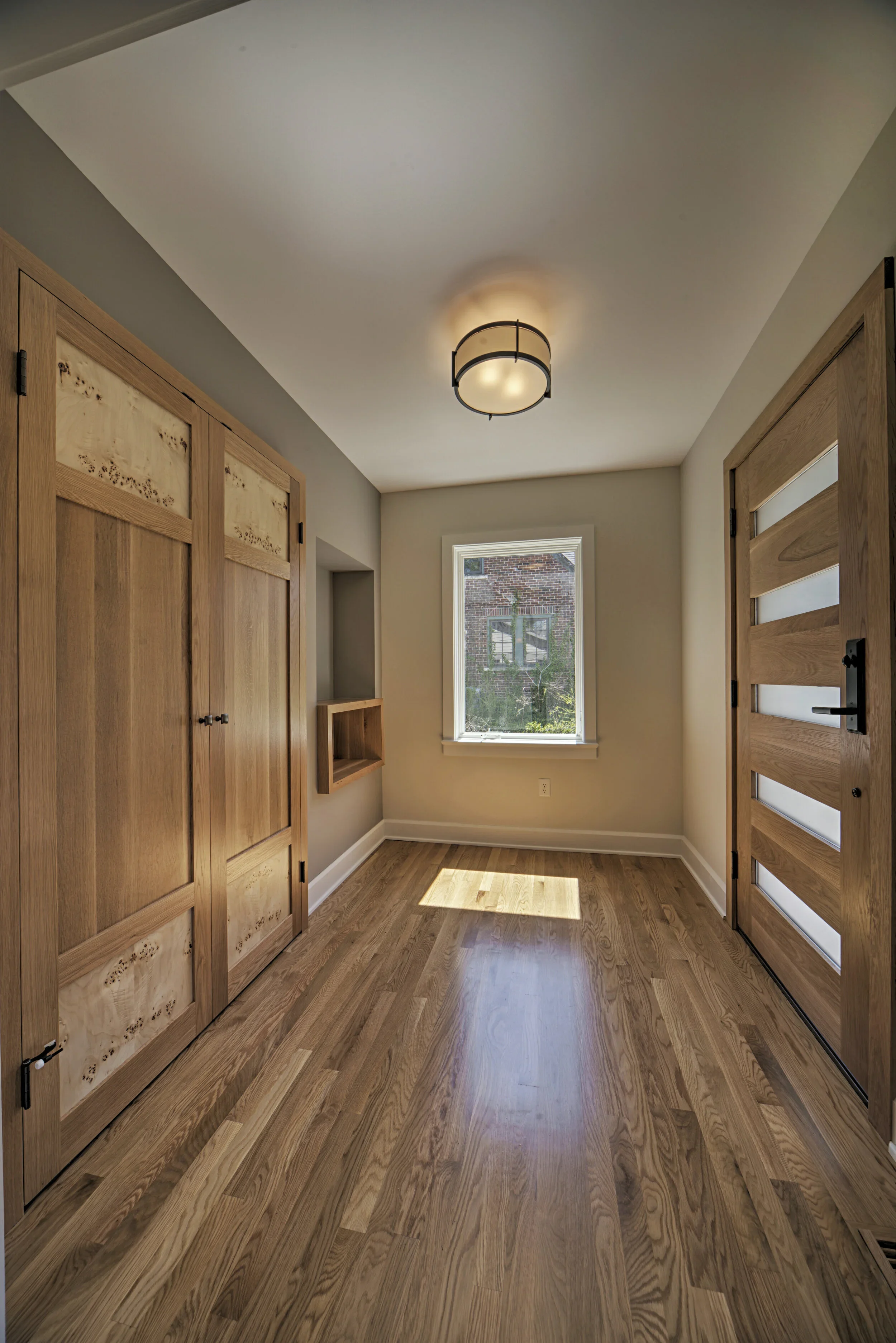 A room with wooden cabinets on the left side, a window at the end, and a modern ceiling light. The floor is wooden, and sunlight is casting a shadow on it.