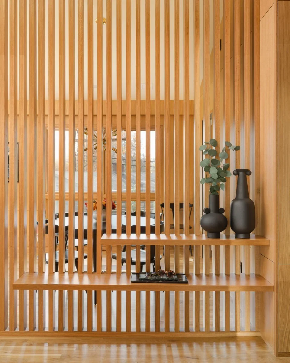 Modern interior with wooden slat divider, overlooking a dining area with a round table and black chairs, large windows with natural light, decorative black vases and a plant on a wooden shelf.