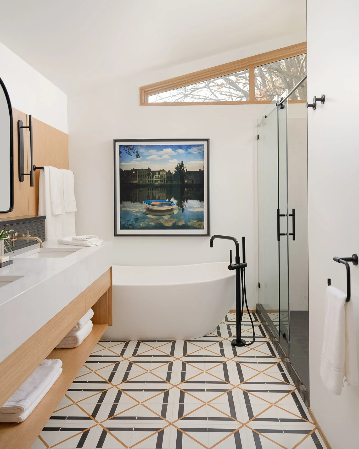 A modern bathroom with a freestanding bathtub, a large framed painting of houses reflected in water, a glass shower with black hardware, a white vanity with wooden shelving and towels, a black towel holder, and a small window with tree branches outsi
