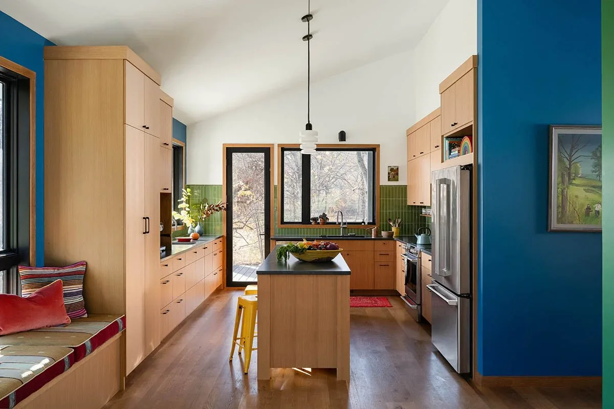 Modern kitchen with wooden cabinets, green tiled backsplash, black countertop, stainless steel appliances, and a central island with yellow stools. Two large windows provide natural light, and there's a colorful gallery wall with artwork.