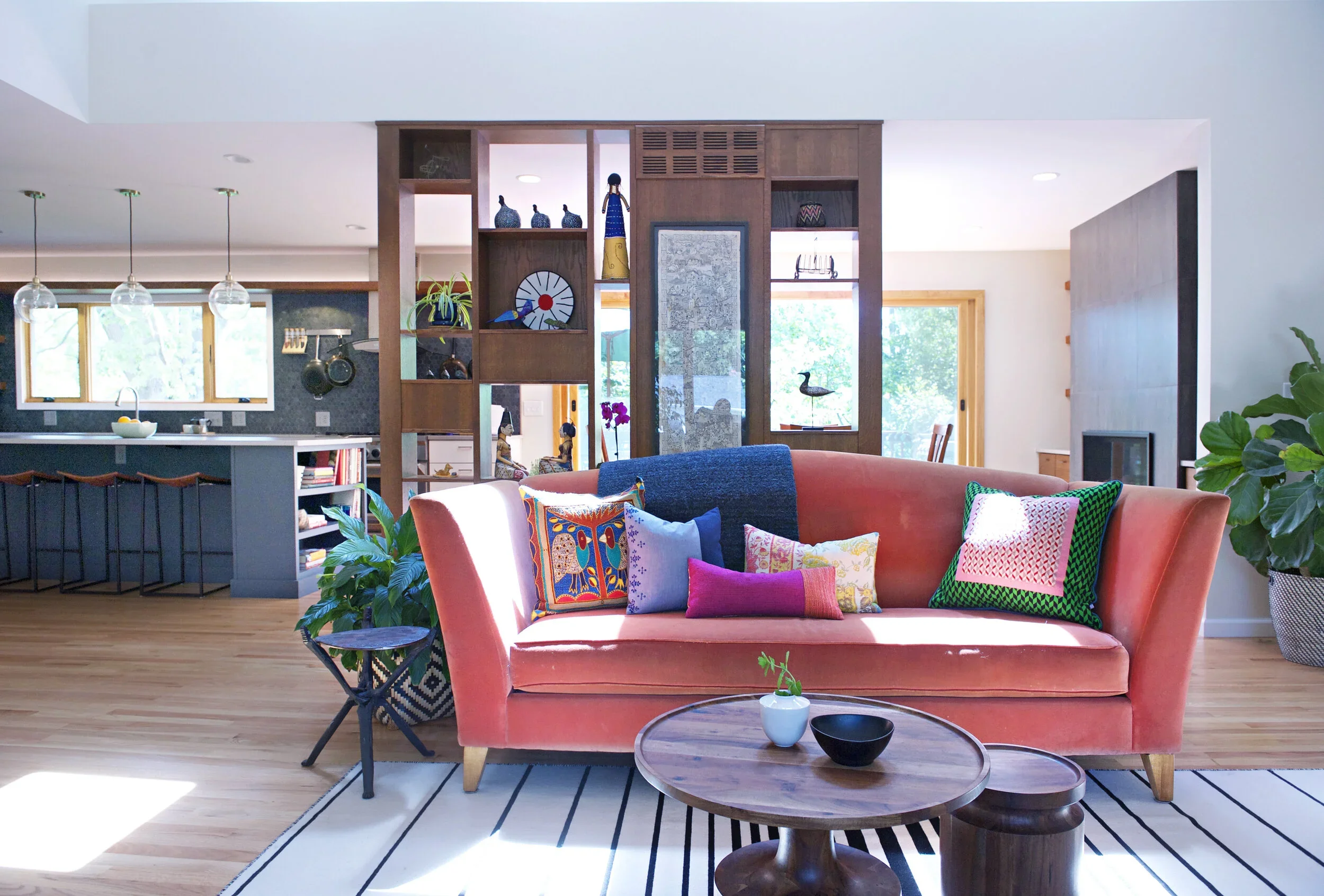 A cozy living room with a pink sofa decorated with colorful patterned pillows, a round wooden coffee table, and a white and black striped rug. There are green plants and natural light coming through large windows, with a wooden partition behind the sofa displaying decorative objects, books, and artwork.