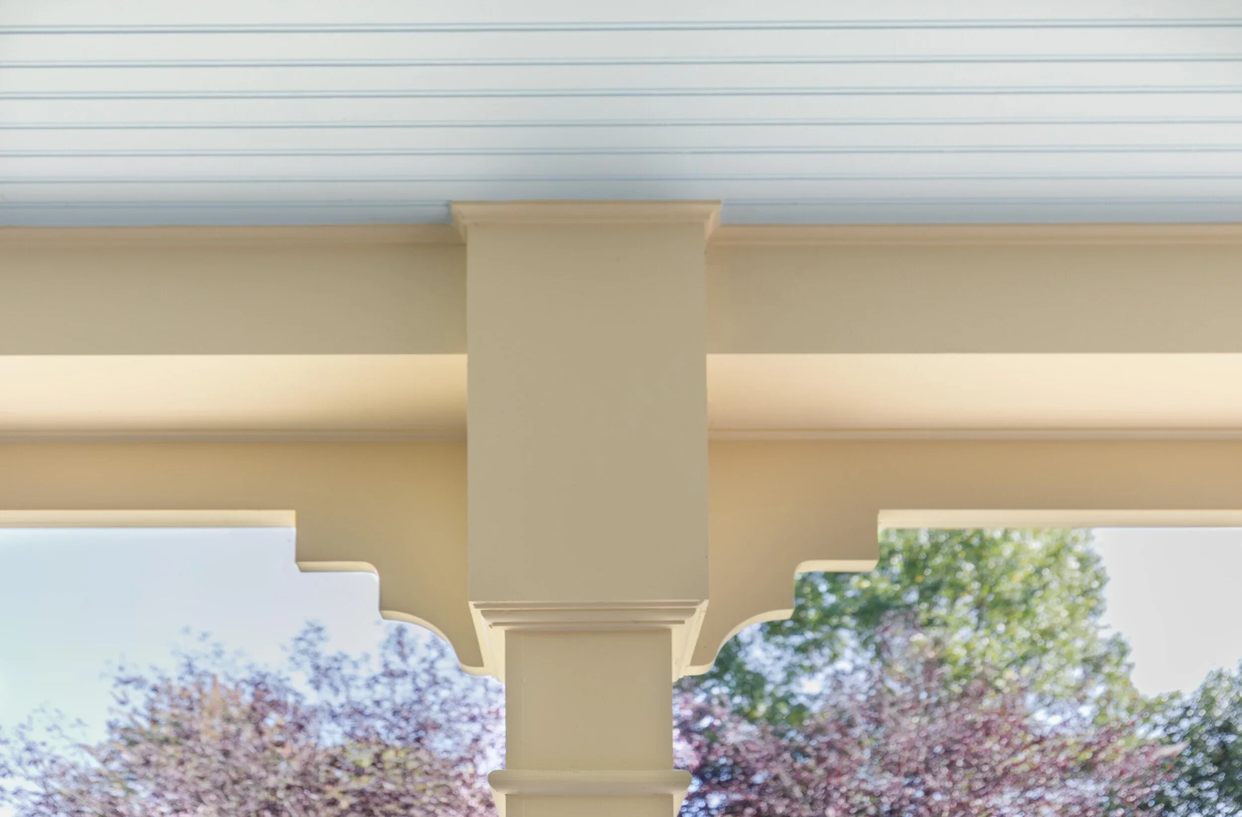 Close-up of white porch columns supporting a roof with horizontal siding, with green trees and blue sky in the background.