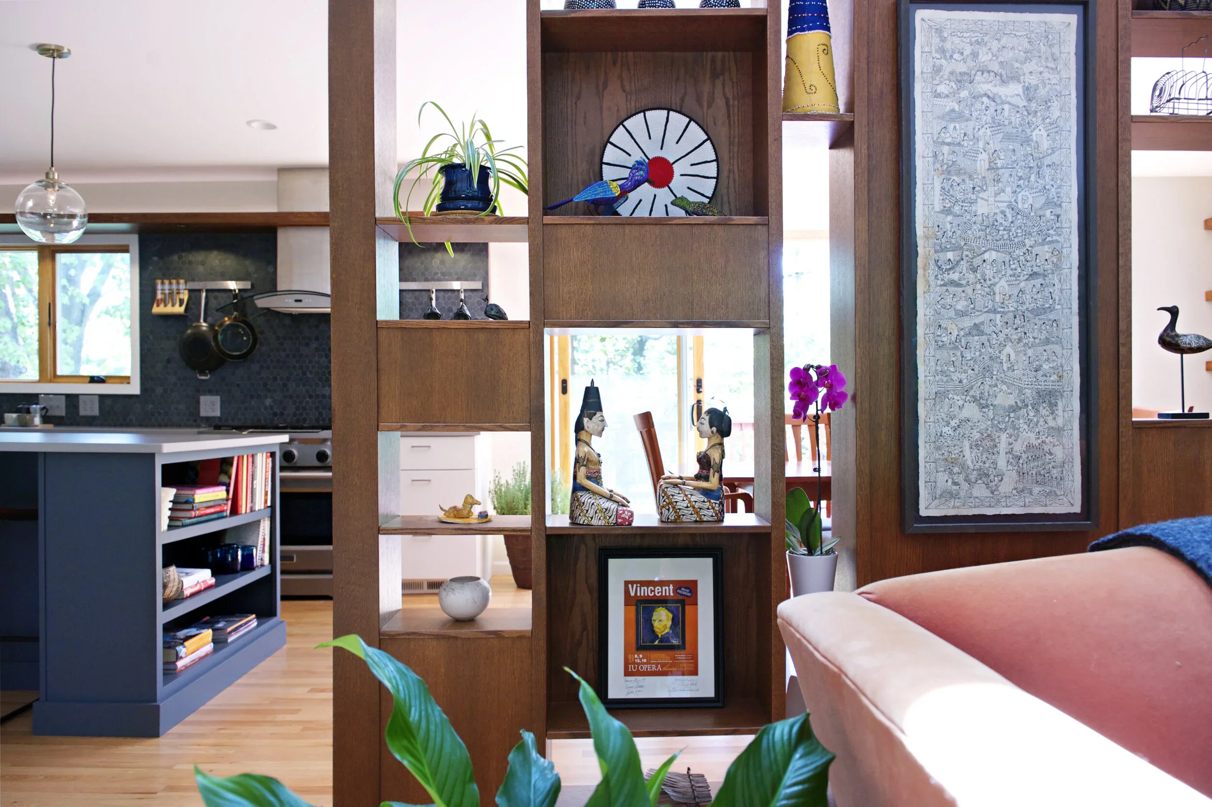 View of a modern living space with decorative wood shelving, artwork, and indoor plants. The background shows a kitchen area with hanging pots, a window, and a dining table with chairs. The room features light wood flooring and contemporary furniture.