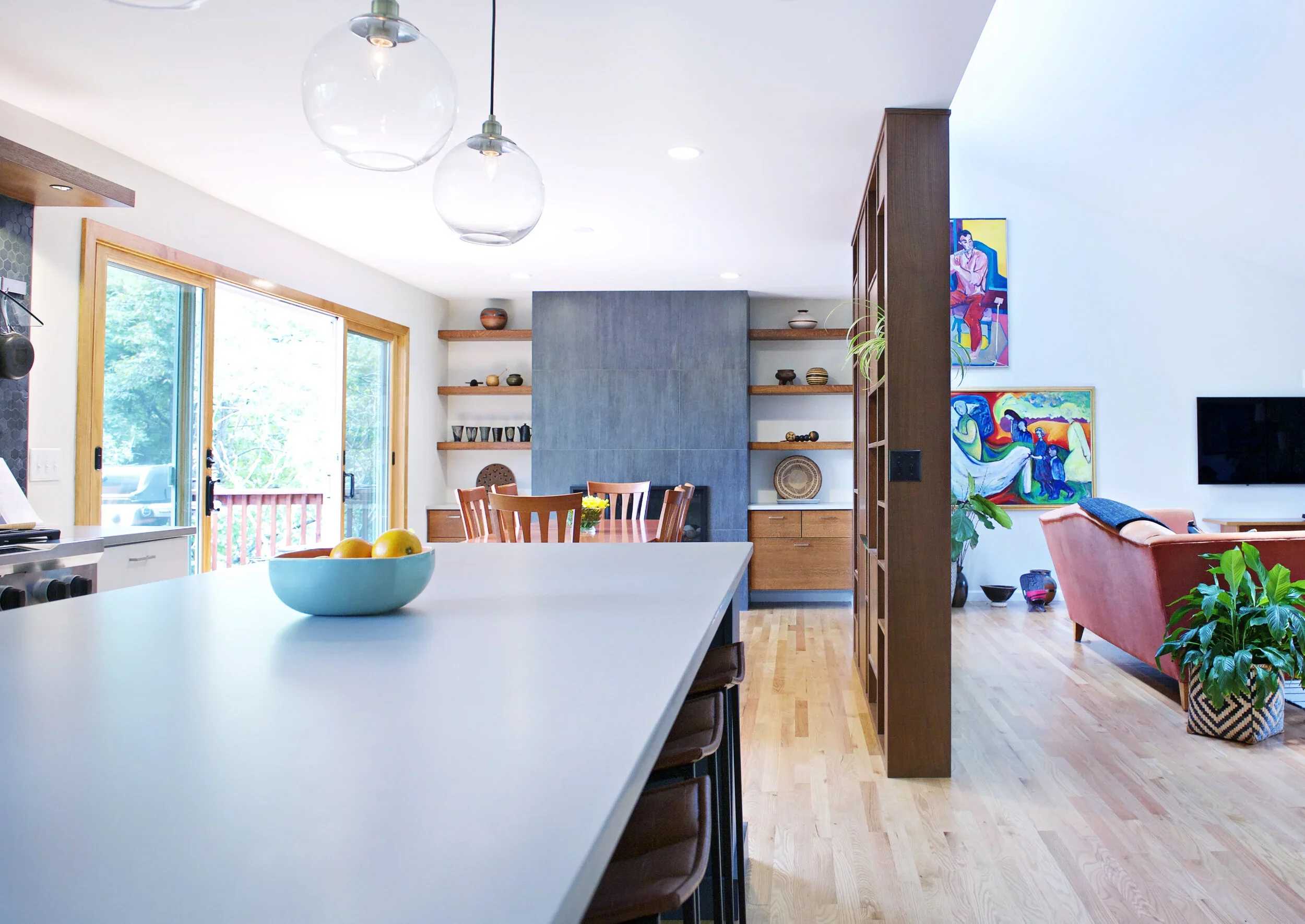 A modern open-concept living and dining area with large windows, wooden floors, colorful artwork on the walls, and a kitchen island with a bowl of yellow apples in the foreground.
