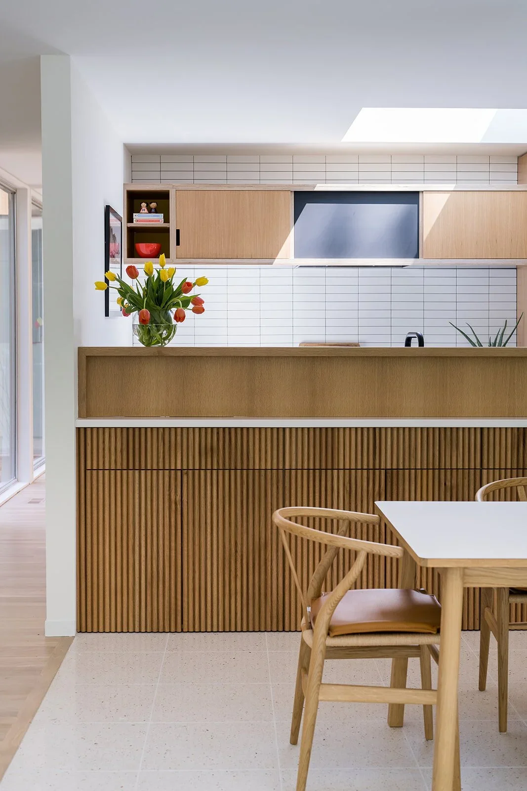 A modern kitchen with wooden cabinetry, white subway tile backsplash, a vase of colorful tulips, and a dining table with chairs.