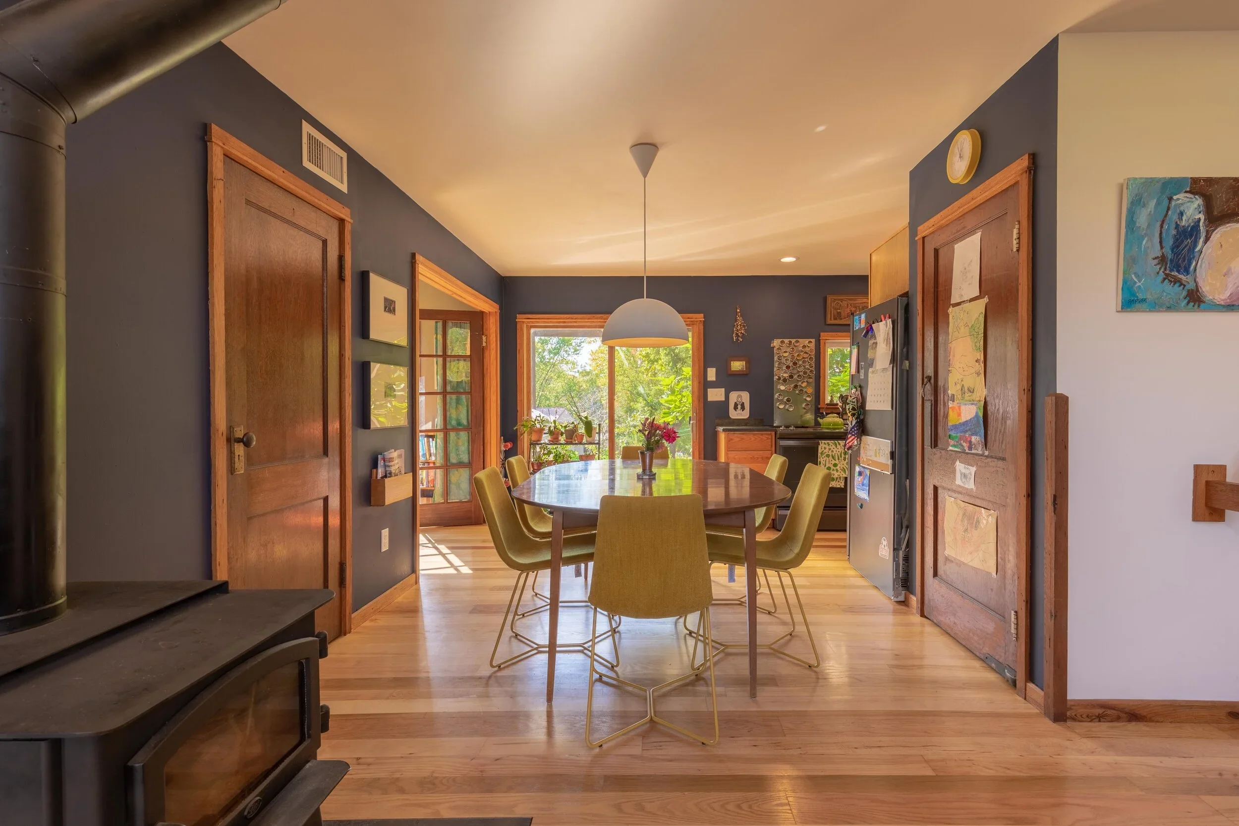 A cozy living and dining area with wooden floors, blue and white walls, and natural light coming through large windows. The room is decorated with artwork, potted plants, and a central round dining table with yellow chairs. A white dome-shaped ceilin