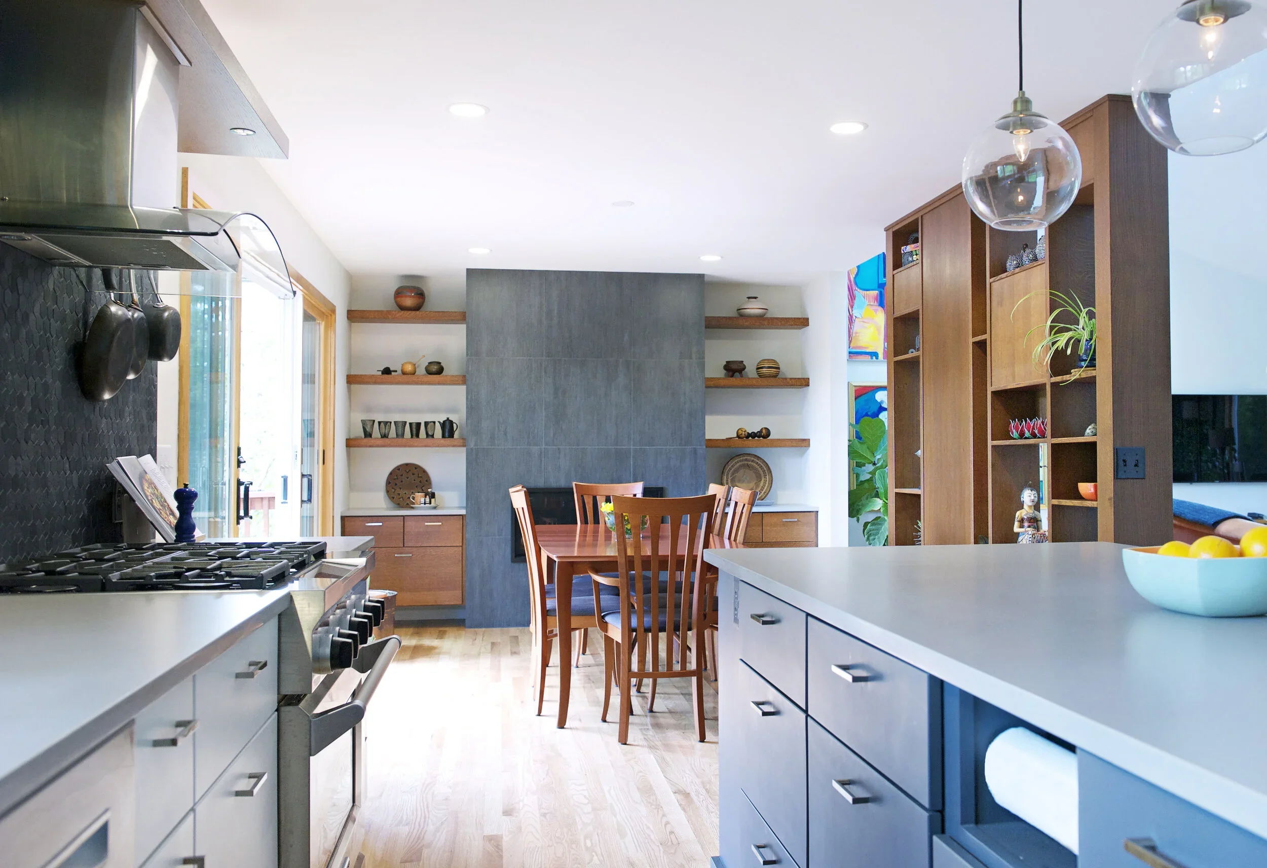 A modern kitchen with white countertops, blue cabinets, and wooden dining table with chairs. Open shelving displays decorative items, and a colorful abstract painting hangs on the wall. Pendant lights and built-in shelving add to the contemporary design.