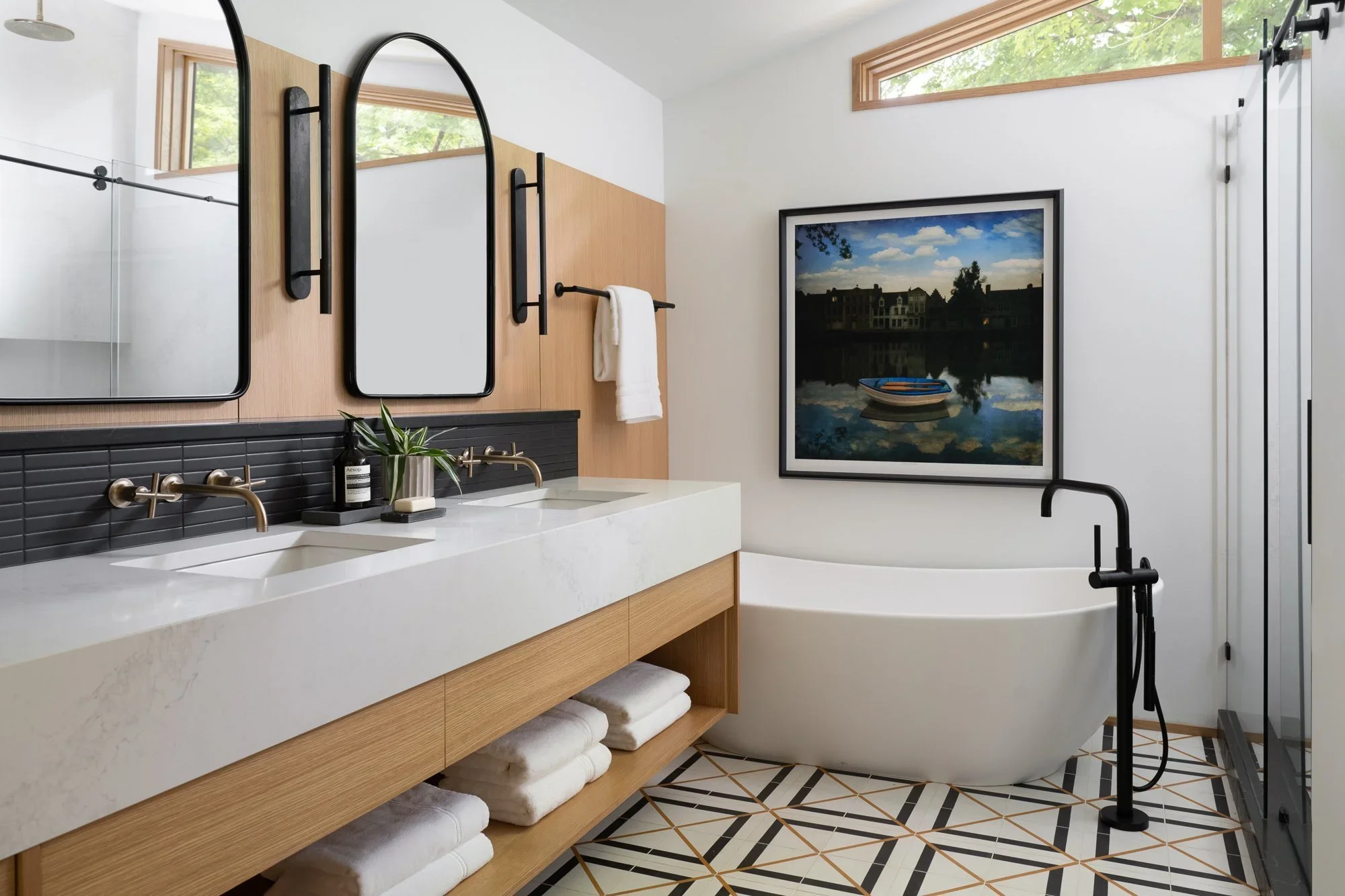 A modern bathroom featuring a double vanity with a concrete countertop, black fixtures, and a small potted plant. There are three mirrors above the vanity and a white bathtub with a black matte shower head and fixtures. The bathroom has a geometric-patterned tile floor, a large framed landscape mirror, and a high horizontal window letting in natural light.