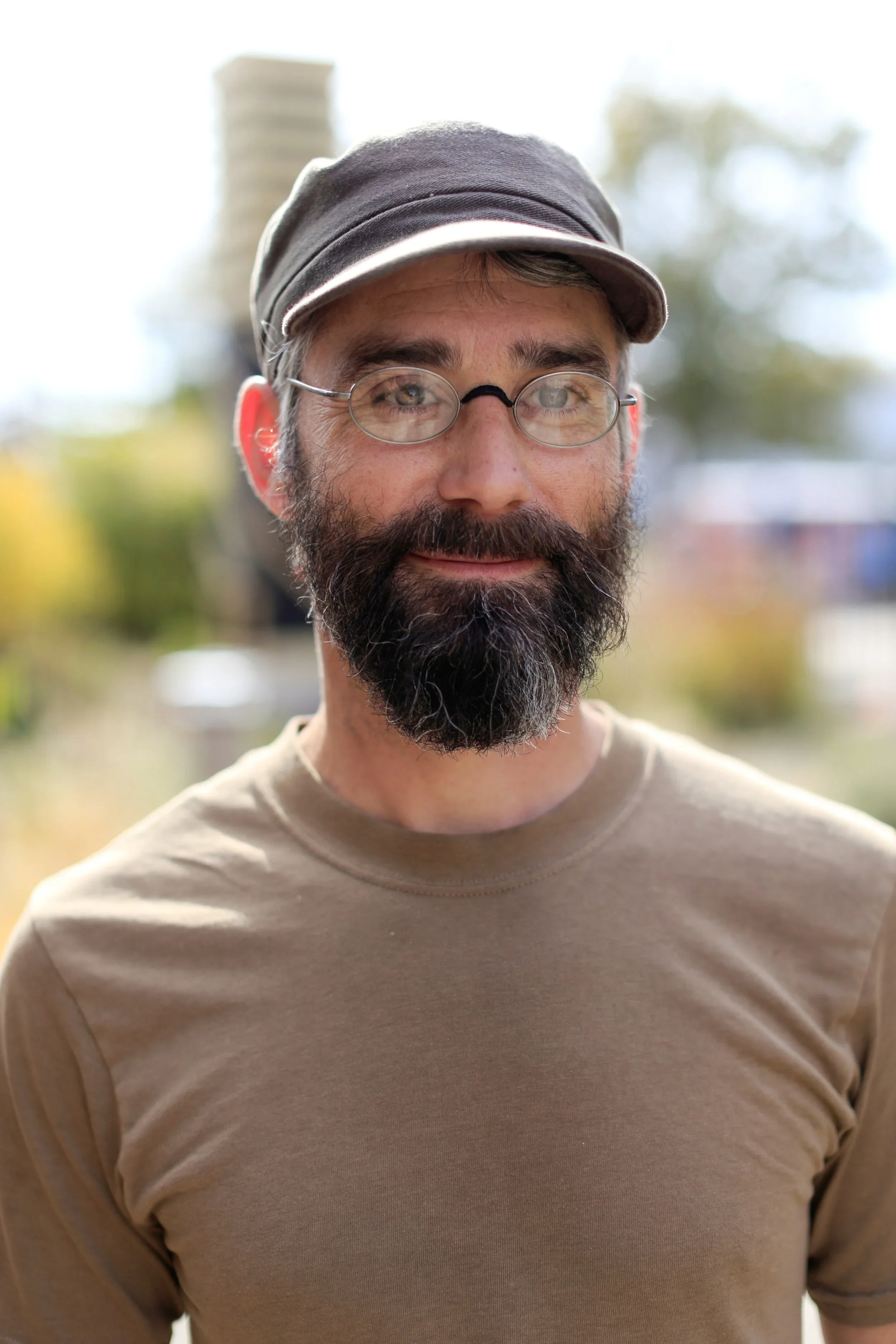 A man with glasses and a beard wearing a cap and a beige shirt outdoors with a blurred background of trees and sky.
