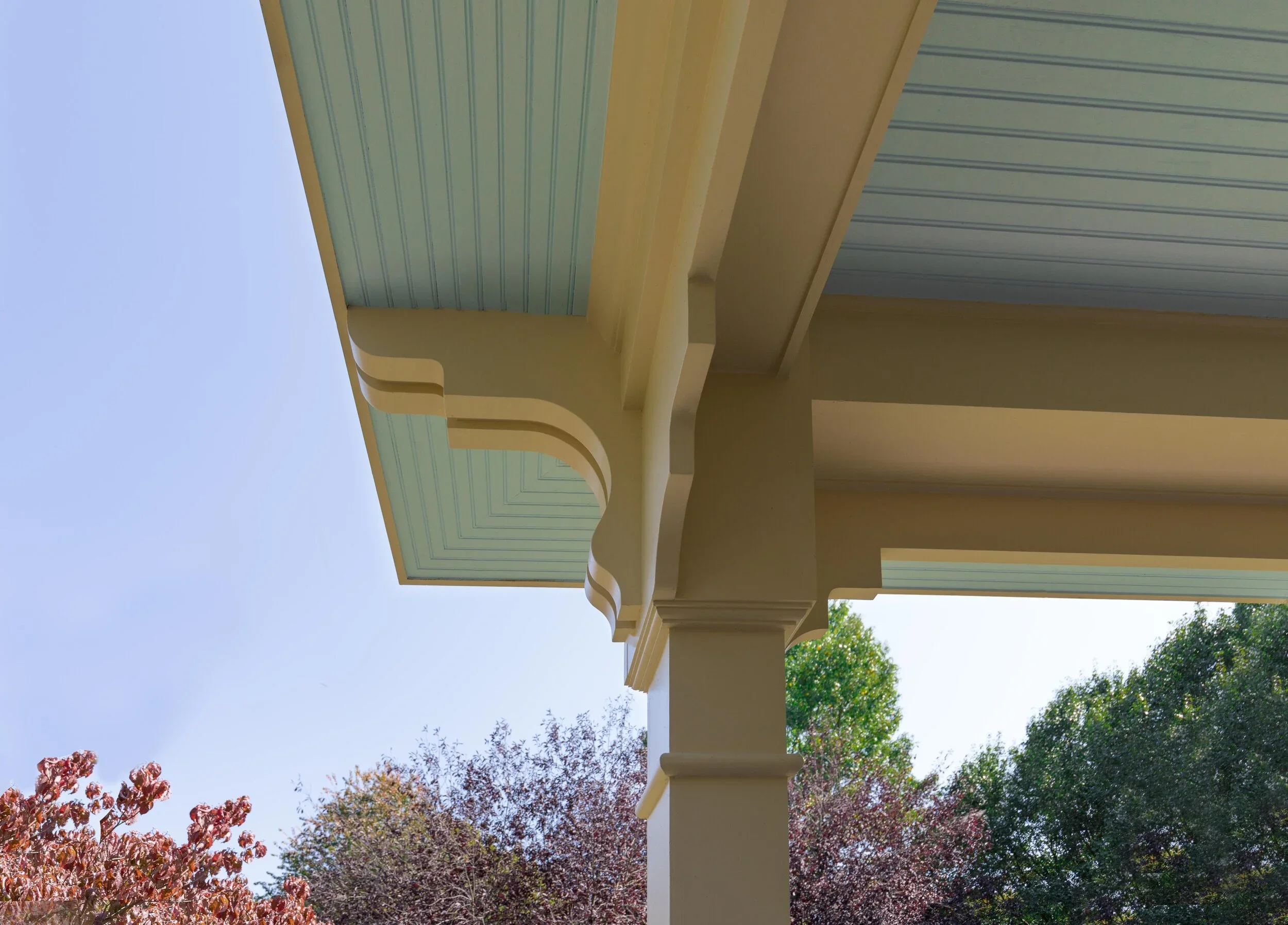 The image shows a section of a porch or balcony with a decorative railing and trim, set against a clear blue sky and trees with autumnal foliage.