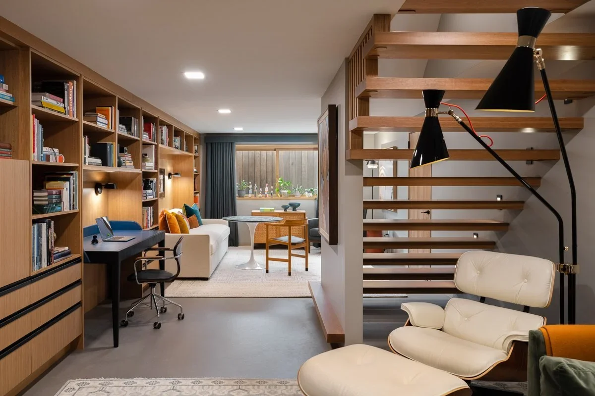 Modern living room with wooden bookshelf, white sofa, round coffee table, desk with chair, large window with curtains, decorative pillows, armchair, unique black floor lamps, and wooden staircase.