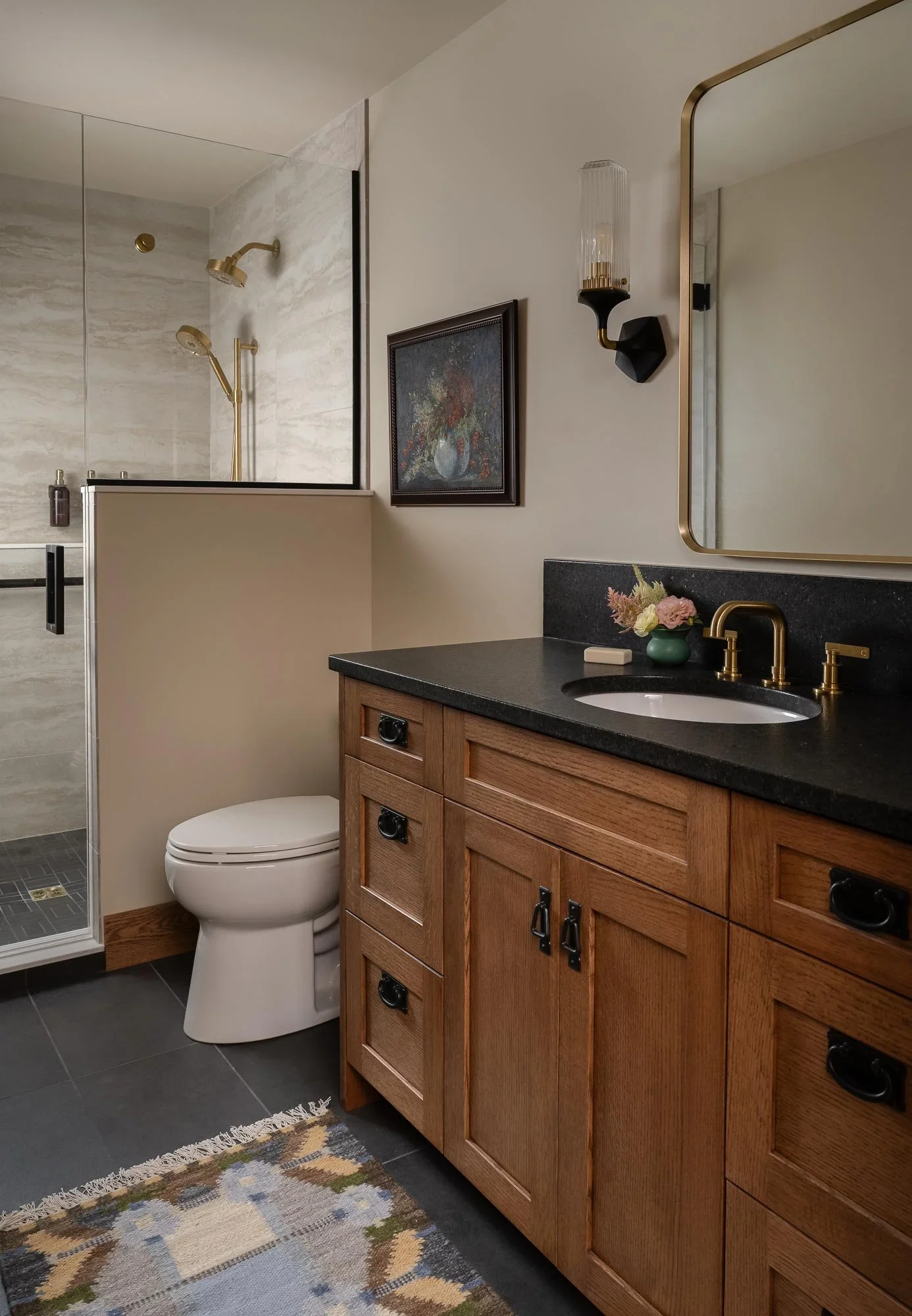 A bathroom featuring a shower with gold fixtures, a white toilet, a wooden vanity with black countertops, a gold faucet, a vase with flowers, a wall mirror with a gold edge, a framed painting, a wall-mounted light, and a patterned rug on dark gray ti