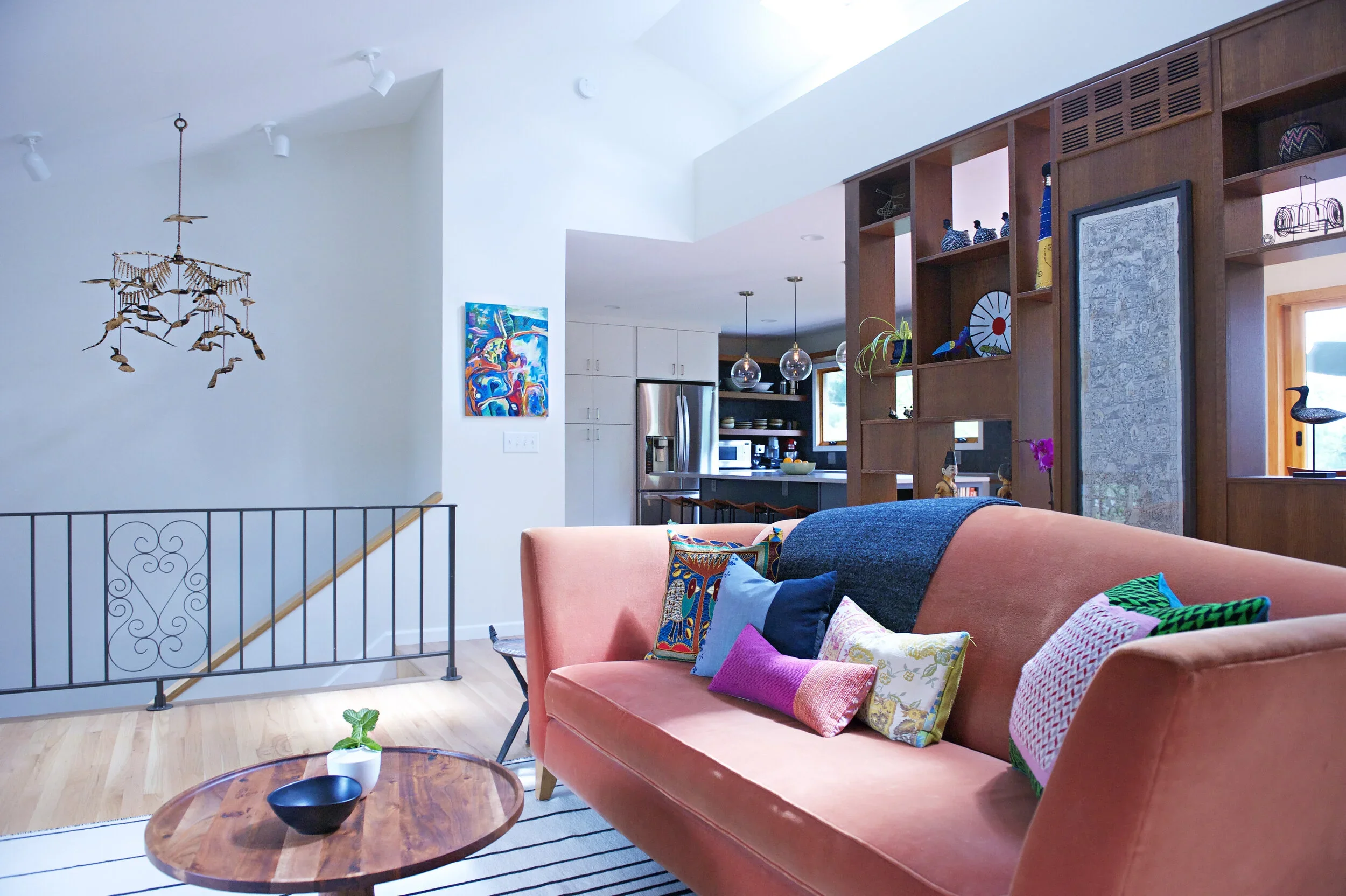 Living room with pink sofa, colorful cushions, wooden shelving unit with decorative items, and a view into the kitchen area with white cabinets and stainless steel refrigerator.