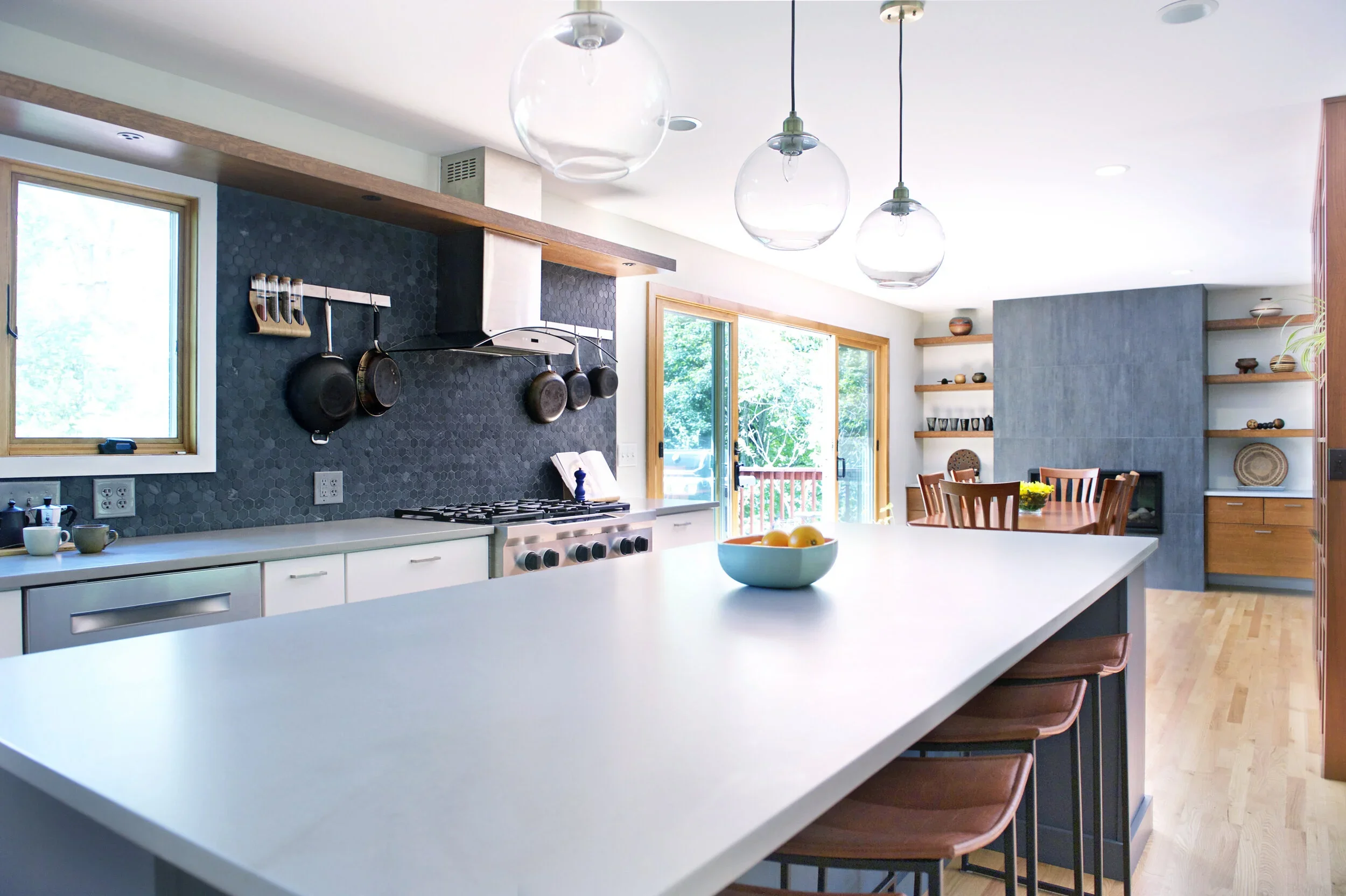 Modern kitchen with white island, wooden chairs, black and gray backsplash, hanging glass pendant lights, and large windows showing green trees outside.