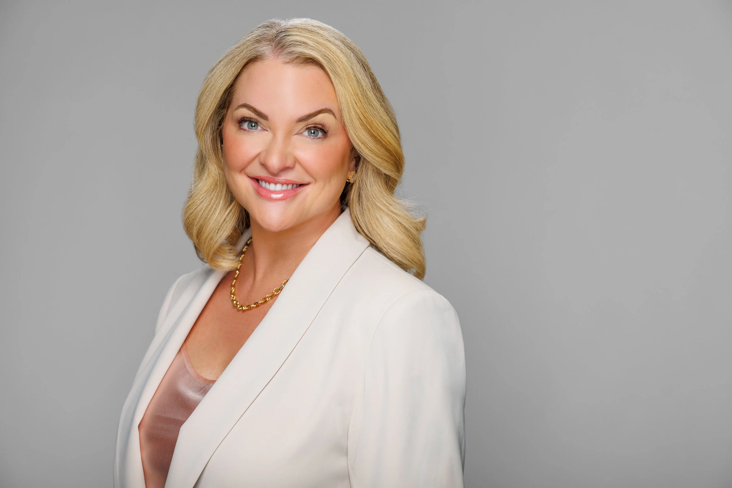 A smiling blonde woman wearing a white blazer and a gold necklace against a gray background.