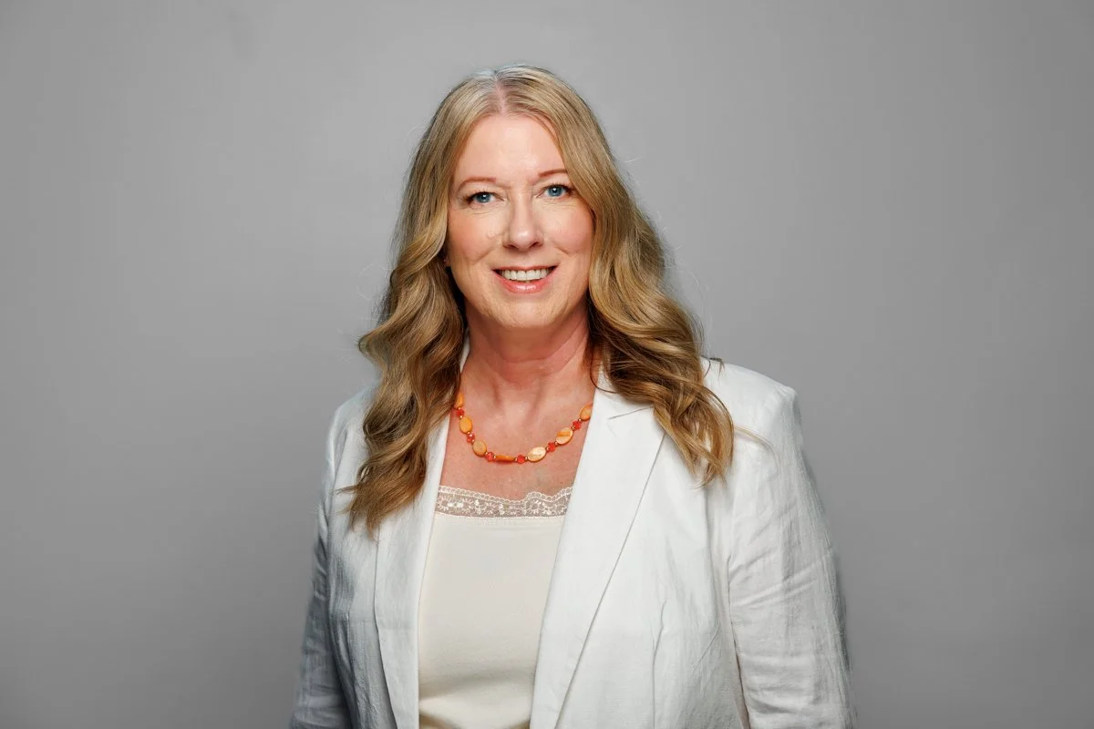 A woman with long, wavy blonde hair wearing a white blazer, a cream-colored top with lace detail, and a colorful beaded necklace.
