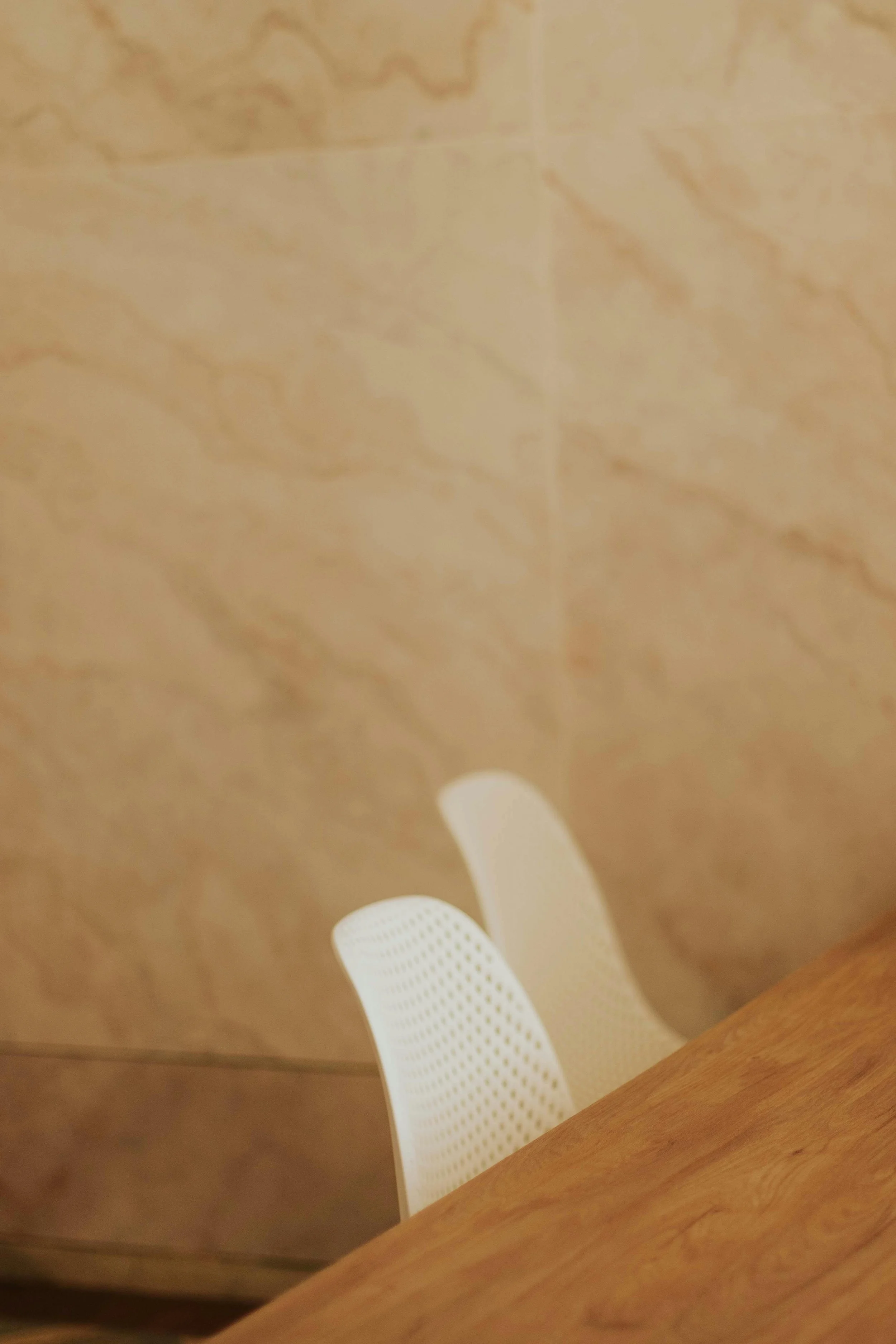 A white plastic chair with a curved backrest and small holes for ventilation, placed at a table with a wooden edge, in a room with beige tiled walls.
