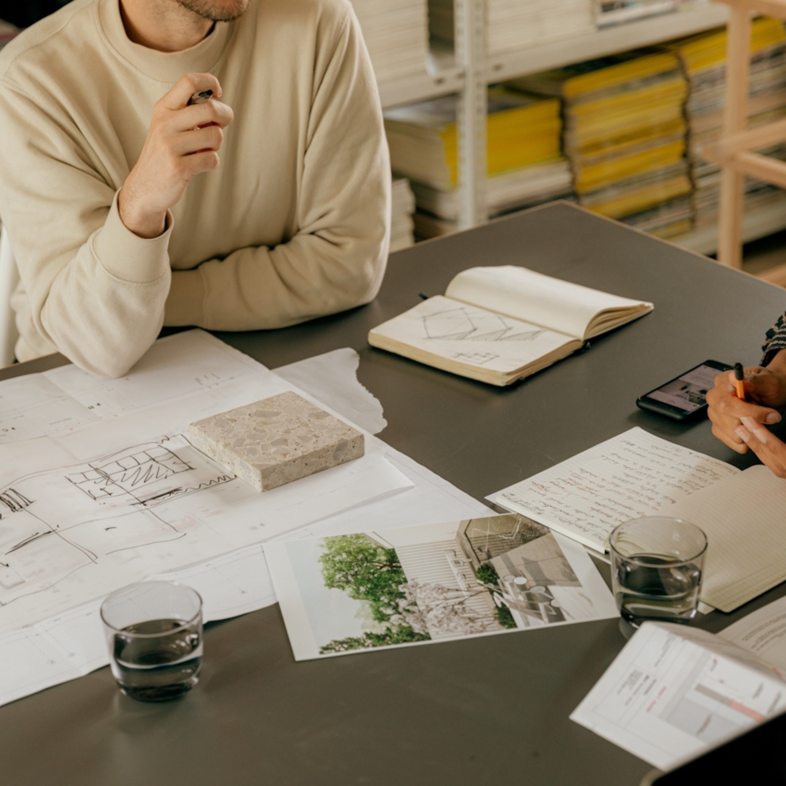 Person in beige hoodie reviewing architecture plans and drawings on a table, with shelves filled with stacks of yellow binders in the background, and glasses of red wine on the table.