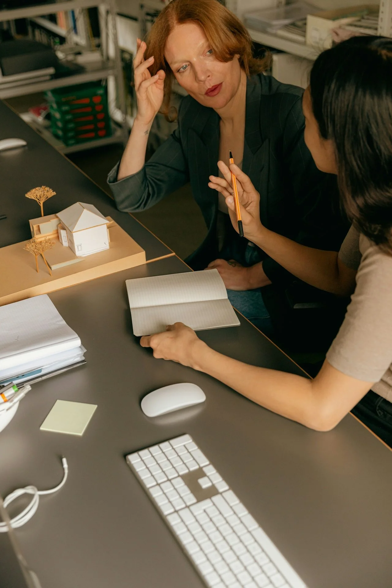 Two women are sitting at a desk in an office, looking at architectural models and discussing design plans.