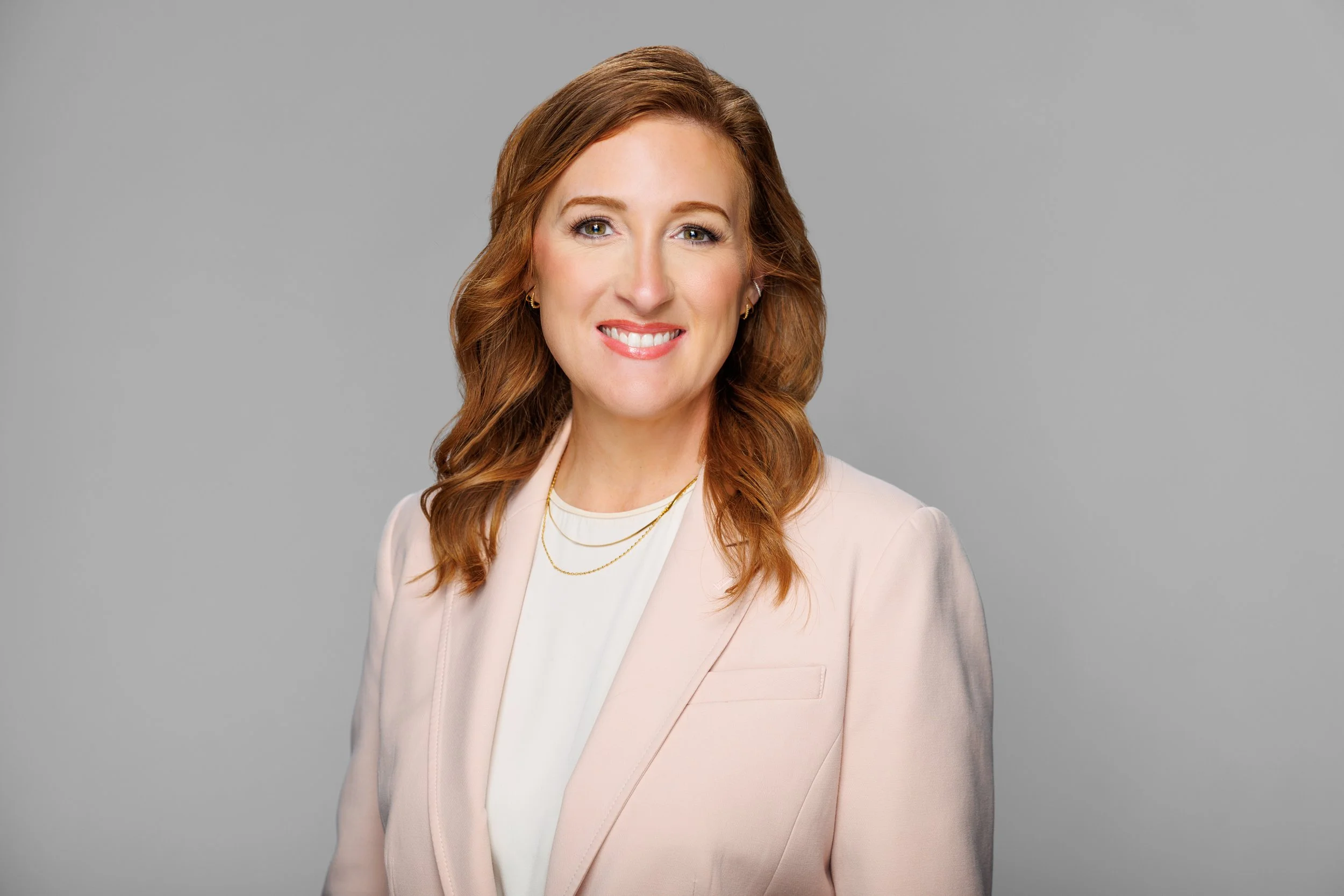 Portrait of a woman with red hair, wearing a light pink blazer over a white top, smiling against a gray background.