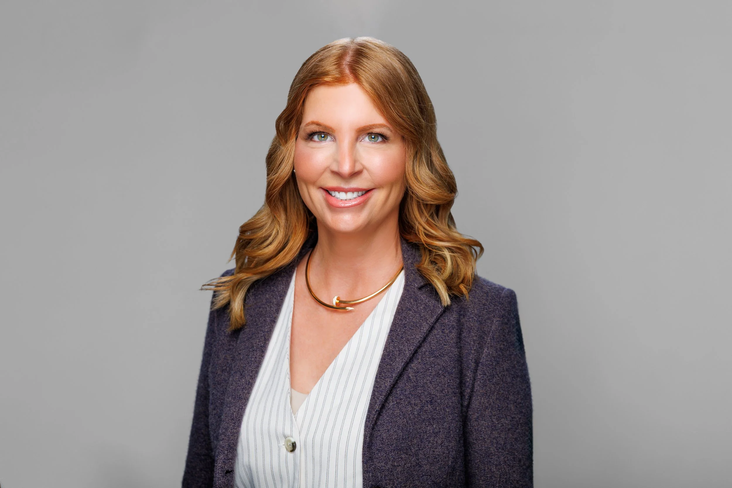 A woman with light skin, shoulder-length wavy red hair, blue eyes, and a friendly smile wearing a striped white blouse, a dark purple blazer, and gold necklaces, standing against a plain gray background.