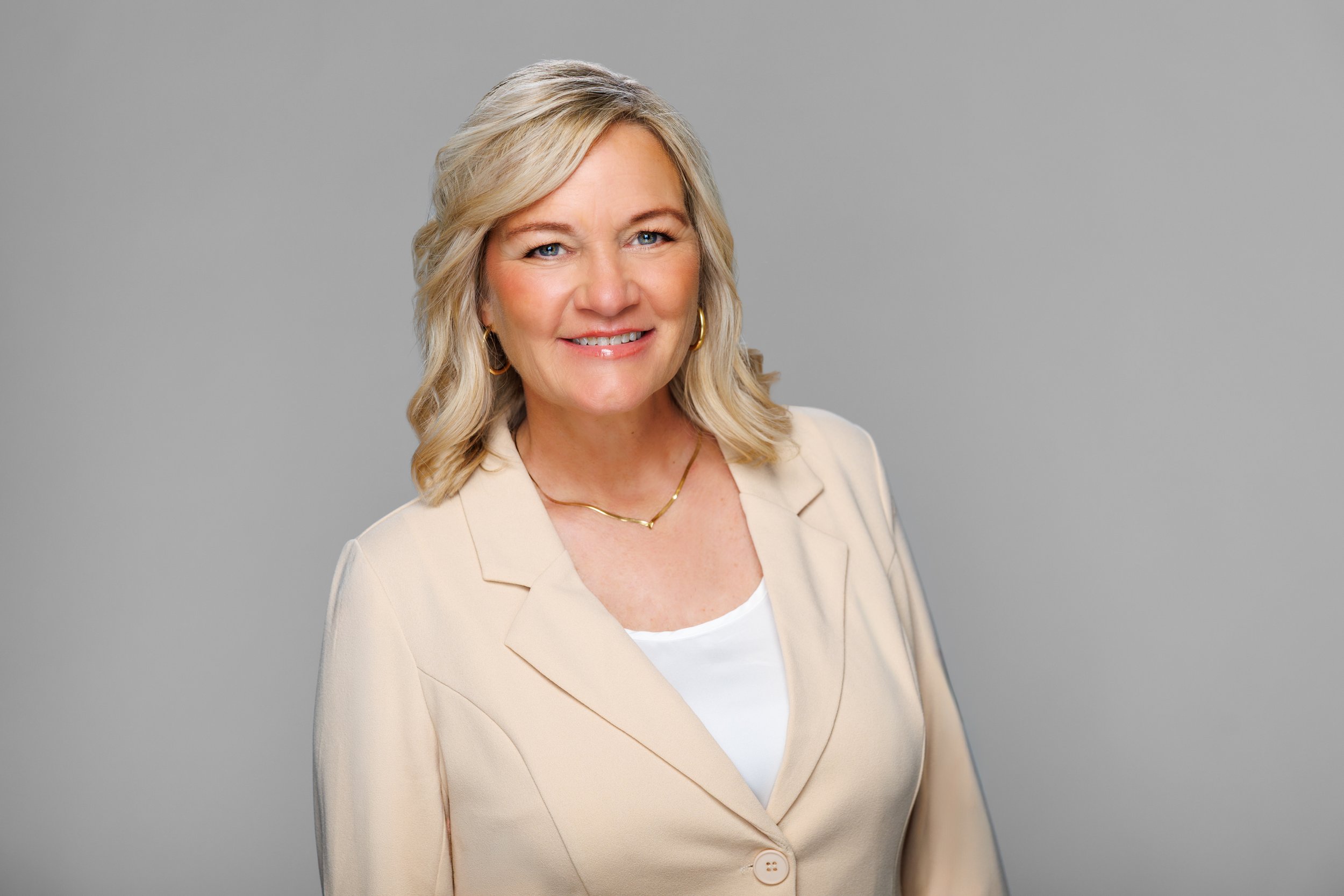 A middle-aged woman with blonde hair styled in loose waves, wearing a beige blazer over a white top, gold hoop earrings, and a delicate gold necklace, standing against a plain gray background and smiling at the camera.