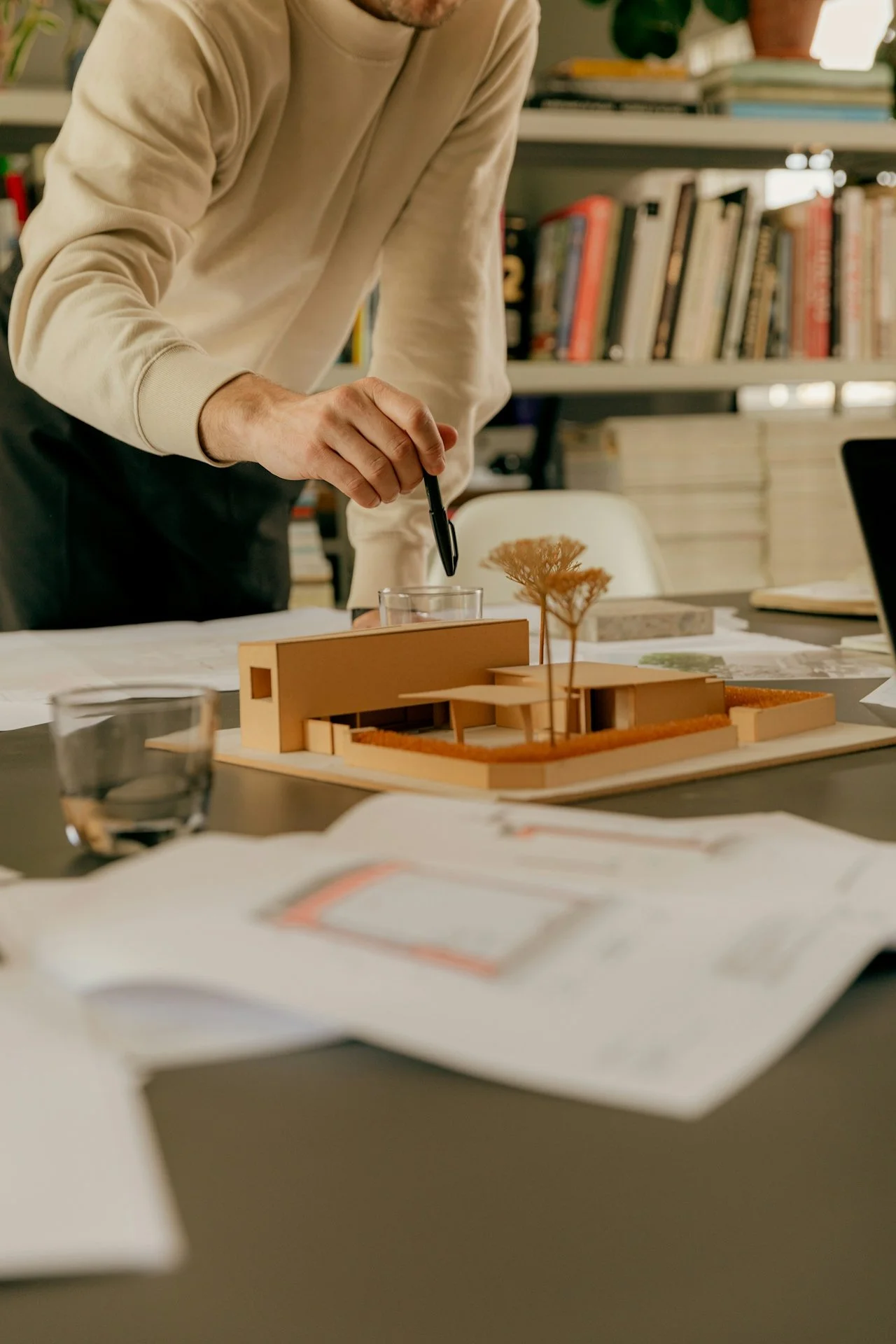 Person working on an architectural model at a table with books and papers in the background.
