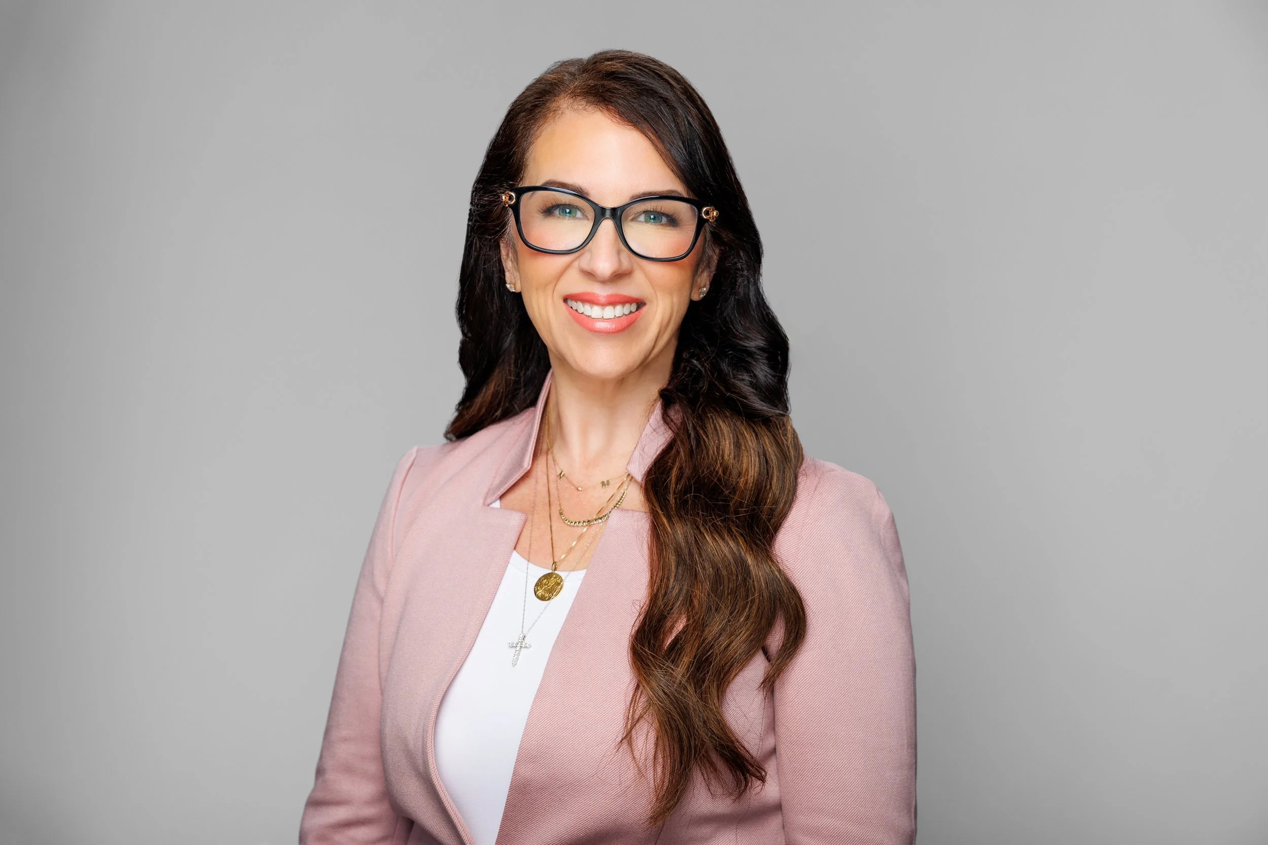 Portrait of a woman with long brown hair, wearing glasses, a pink blazer, a white top, and gold jewelry, smiling against a gray background.